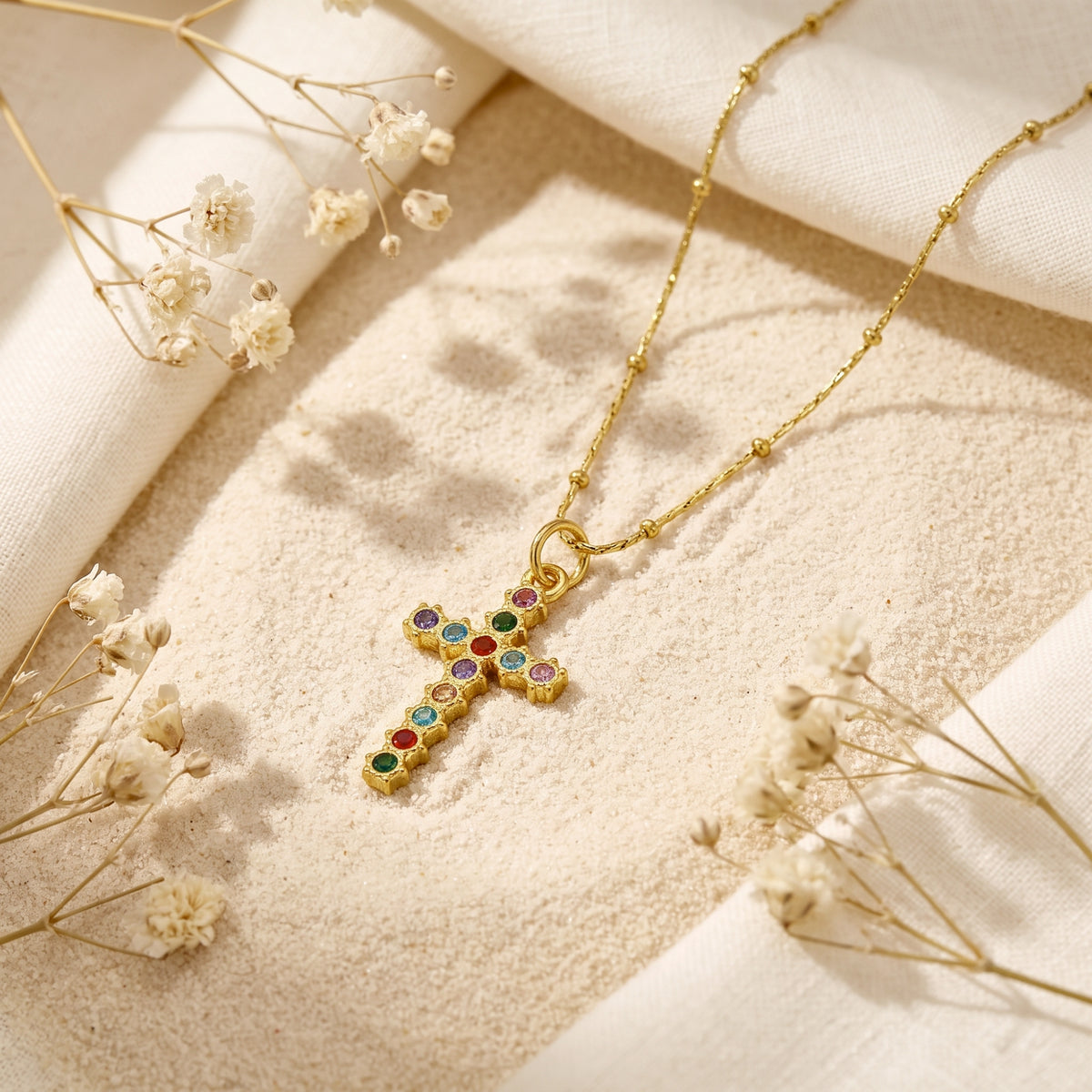 Gold-tone cross pendant with colorful stones resting on pale sand among dried white flowers.