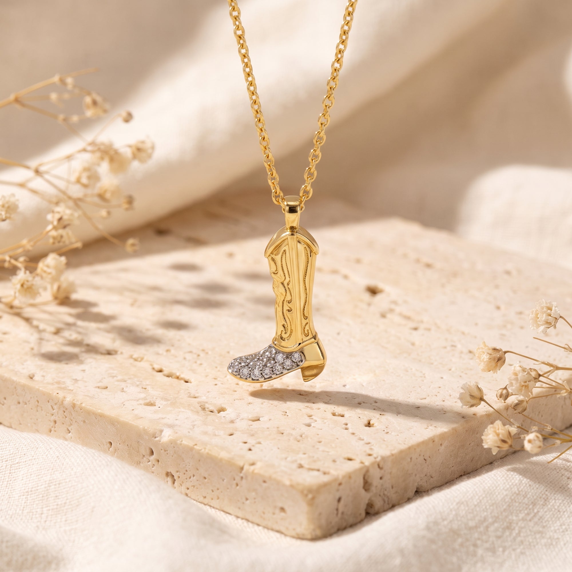 Gold-tone cowboy boot pendant necklace with clear pave stones on the toe, hanging over a beige stone block.
