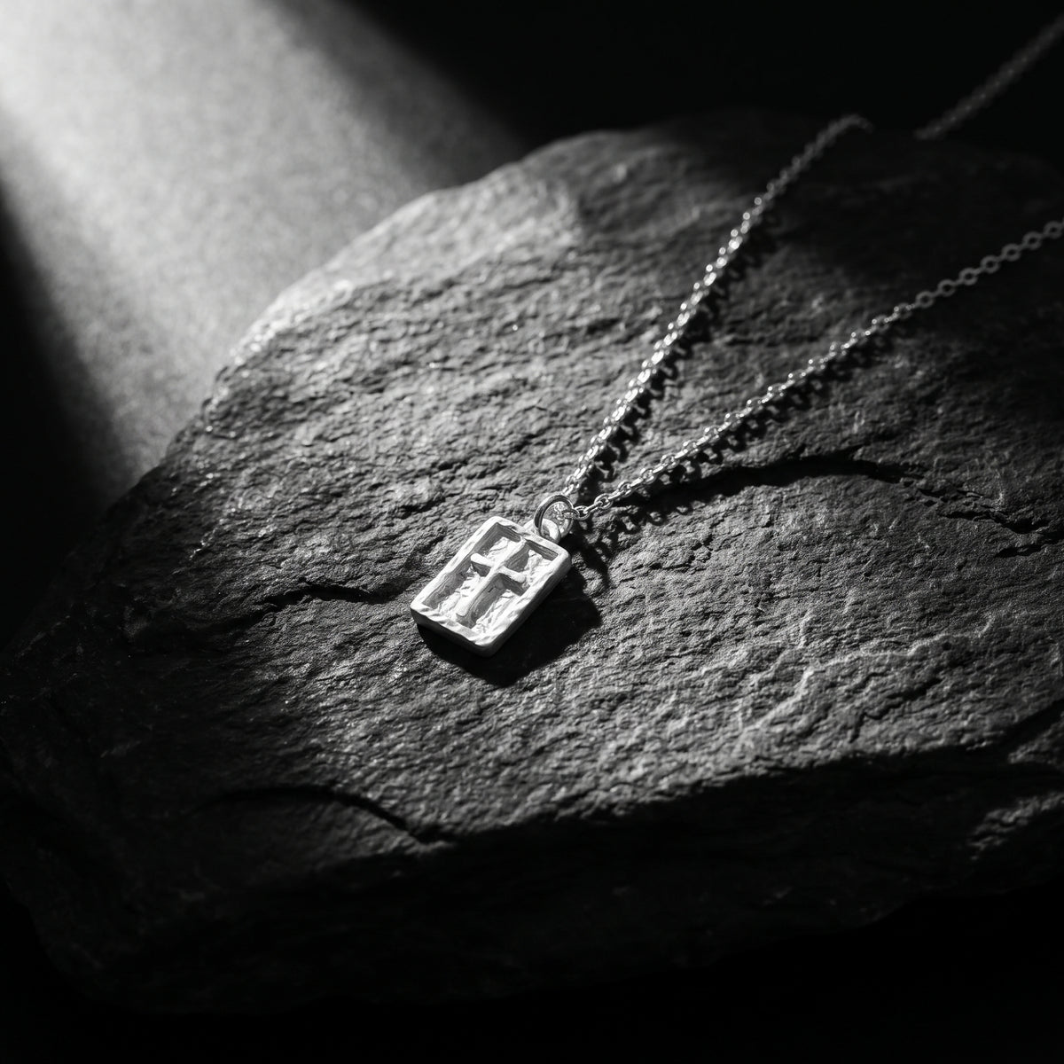 Textured silver rectangular cross pendant laying flat on dark slate stone under dramatic light.