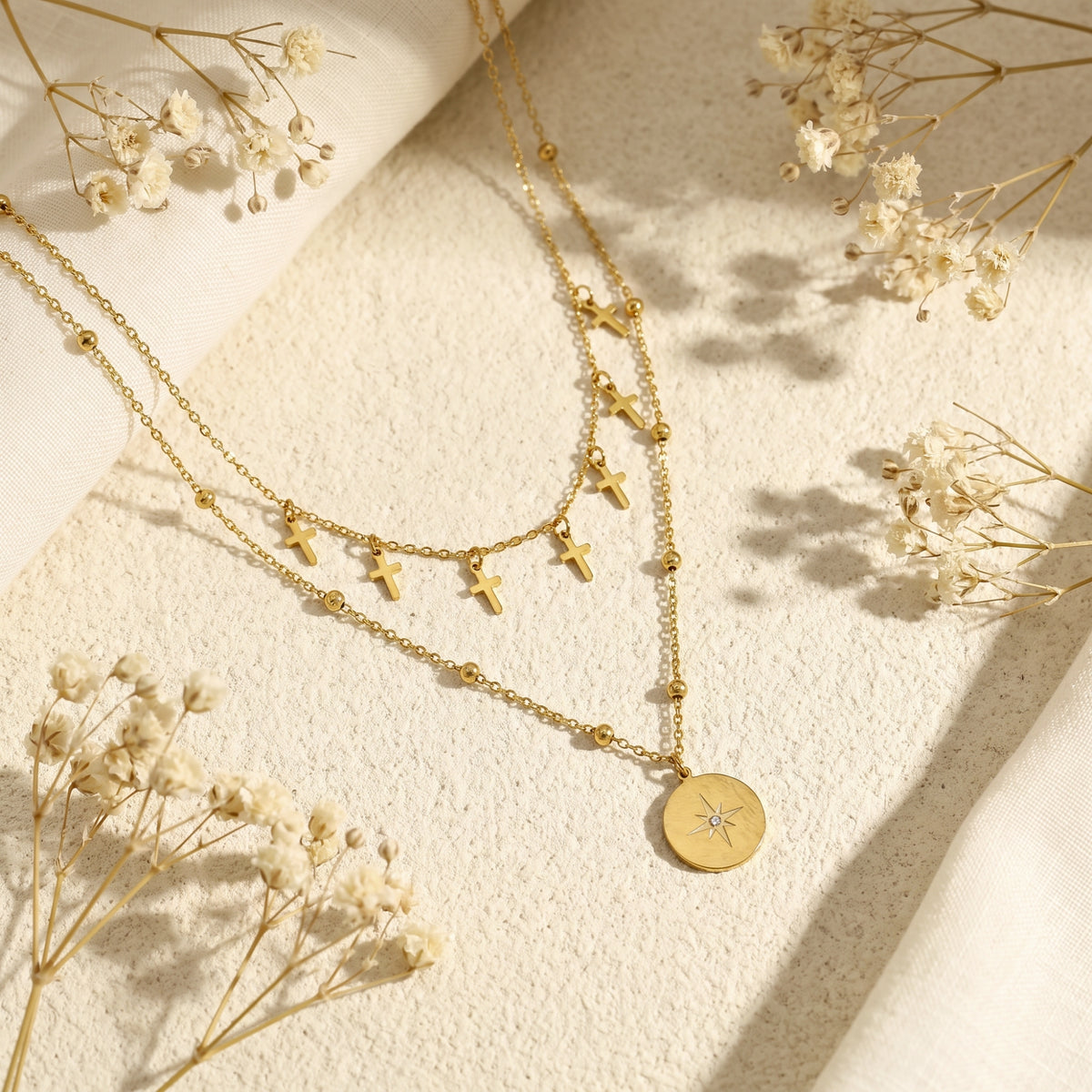 Flat lay of a gold-tone layered necklace with cross charms and star medallion pendant among dried flowers.