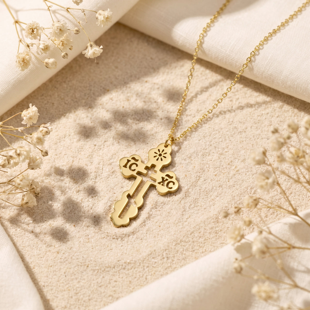 Gold-tone scalloped Orthodox cross pendant and chain resting flat on light sand with white dried flowers.