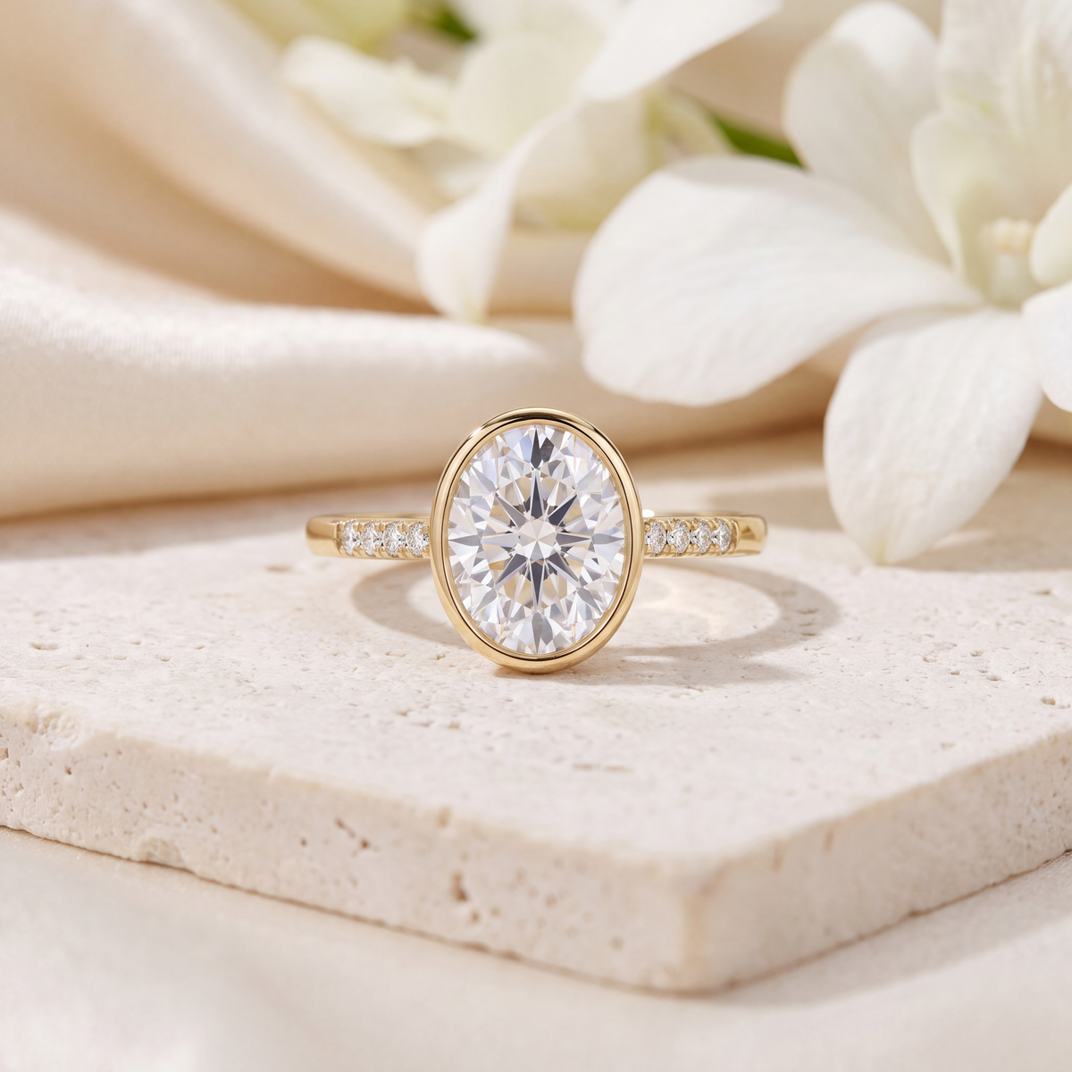 Yellow gold oval moissanite bezel ring with pavé accent stones set on a beige stone surface.