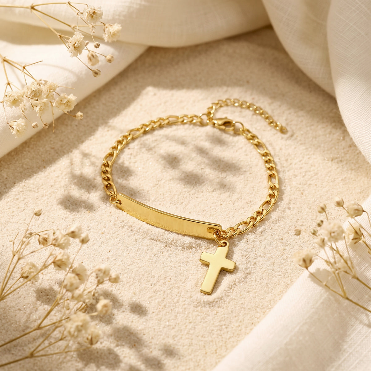 Gold-tone Figaro chain bracelet with blank ID bar and dangling cross charm displayed on sand with dried flowers.