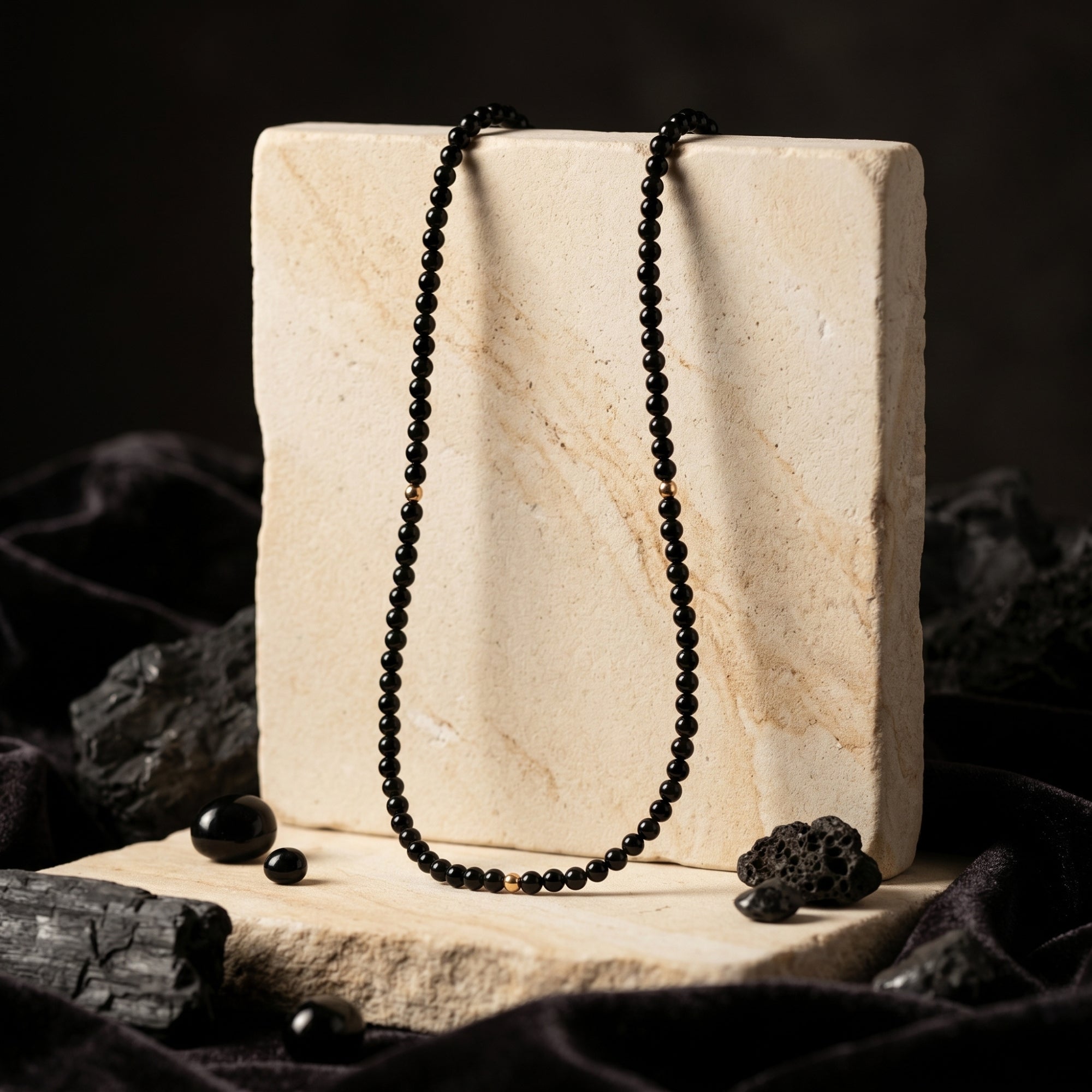 3mm polished black onyx bead necklace with 18K gold accents draped over a stone block in a moody lifestyle setting.