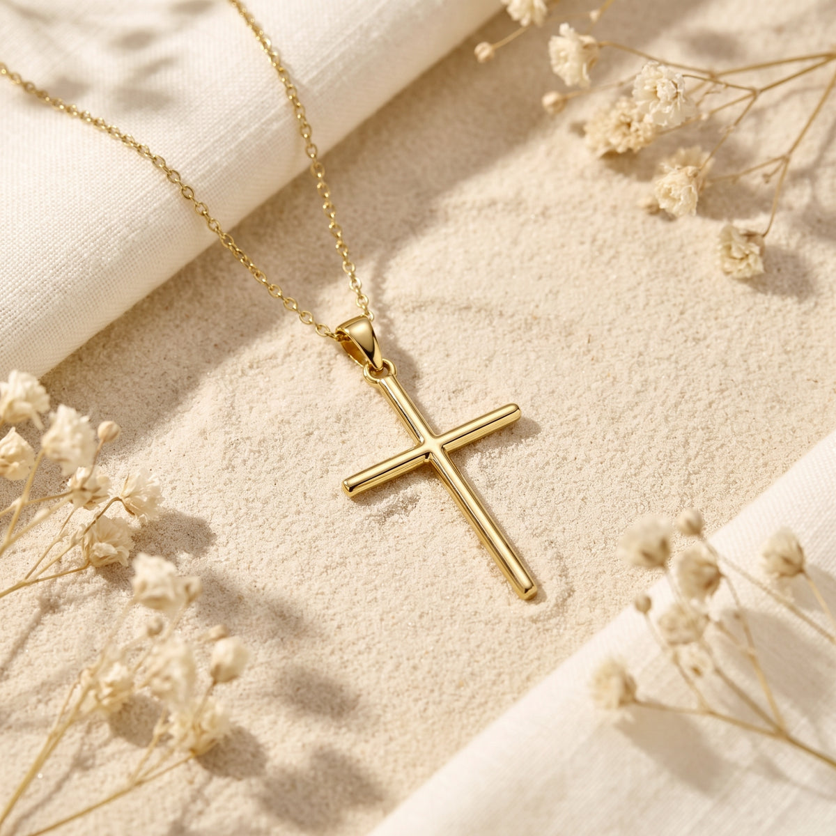 A gold-tone cross necklace lying on fine sand with white cloth and dried baby's breath flowers.