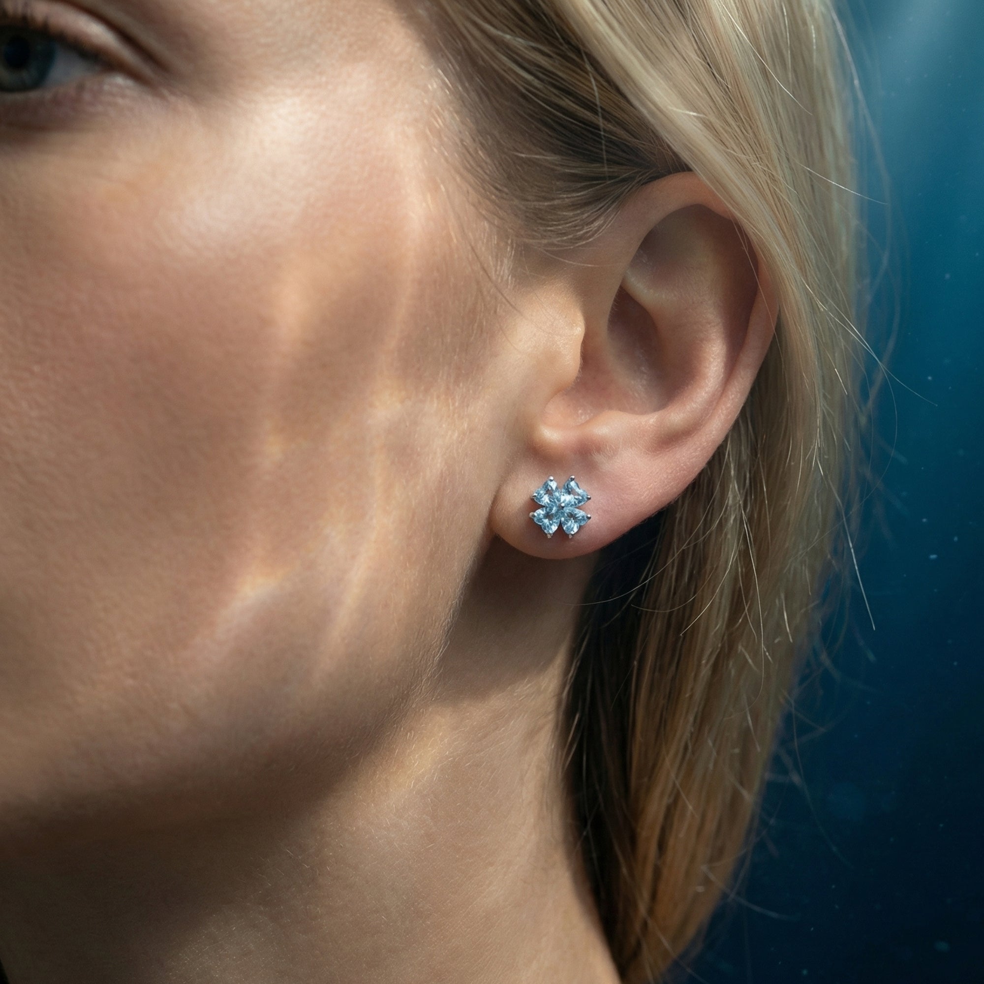 Model wearing a platinum clover stud earring with heart-cut aquamarine stones under dappled light.
