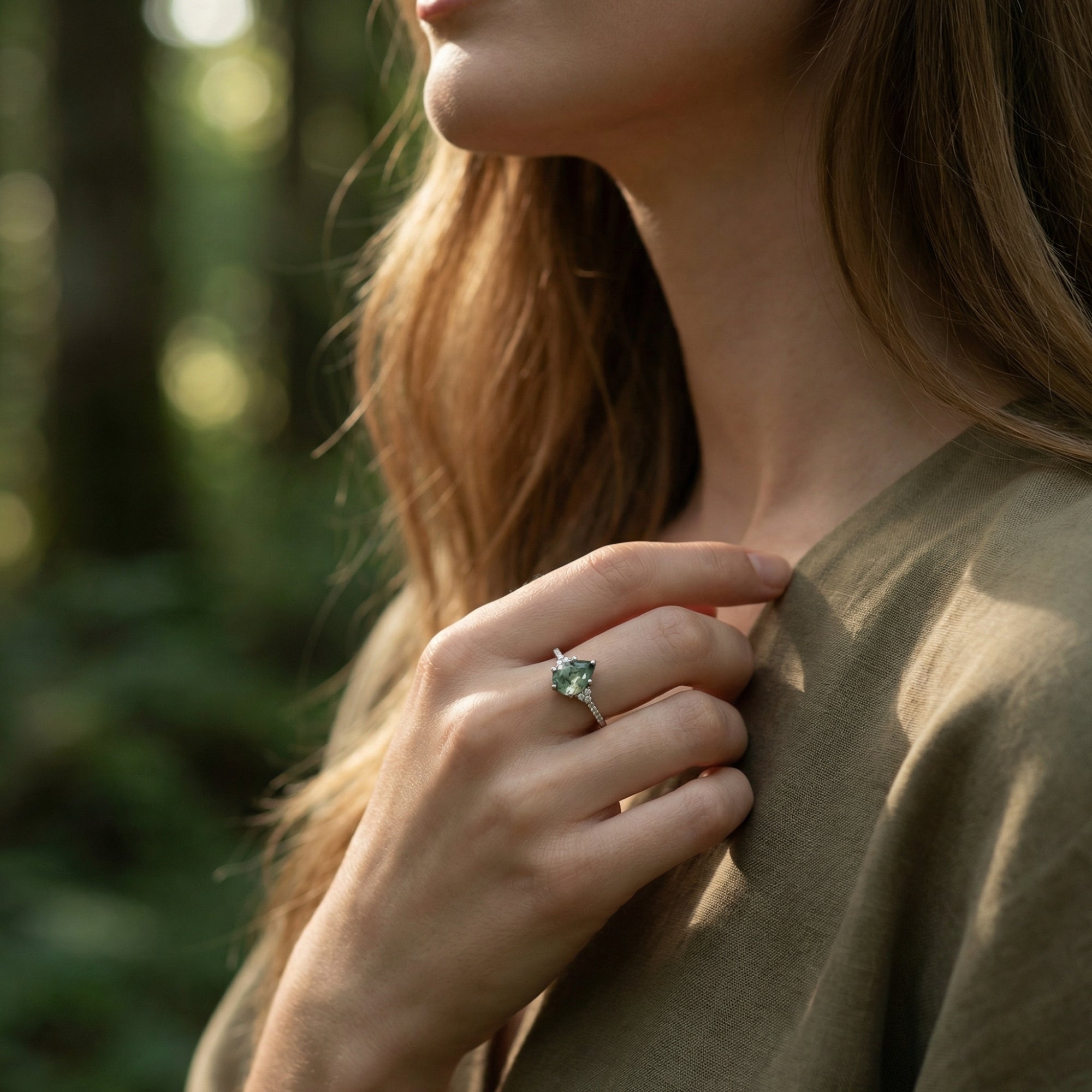 Model wearing a pear cut moss agate ring on her finger in a sunlit forest setting.
