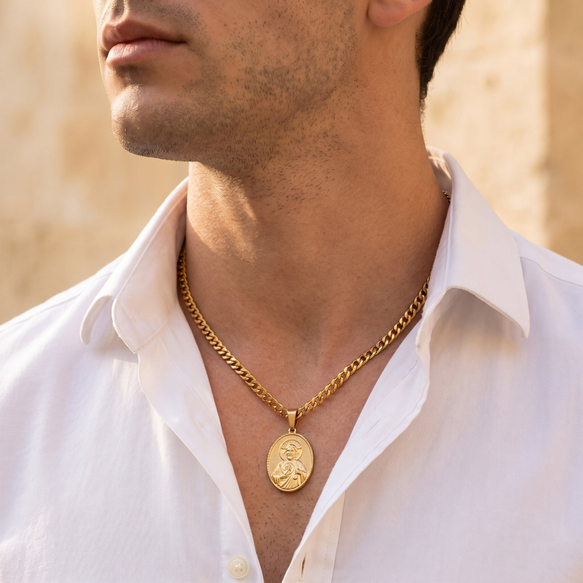 Man wearing a gold-tone oval Saint Jude medal necklace over an open white collared shirt.