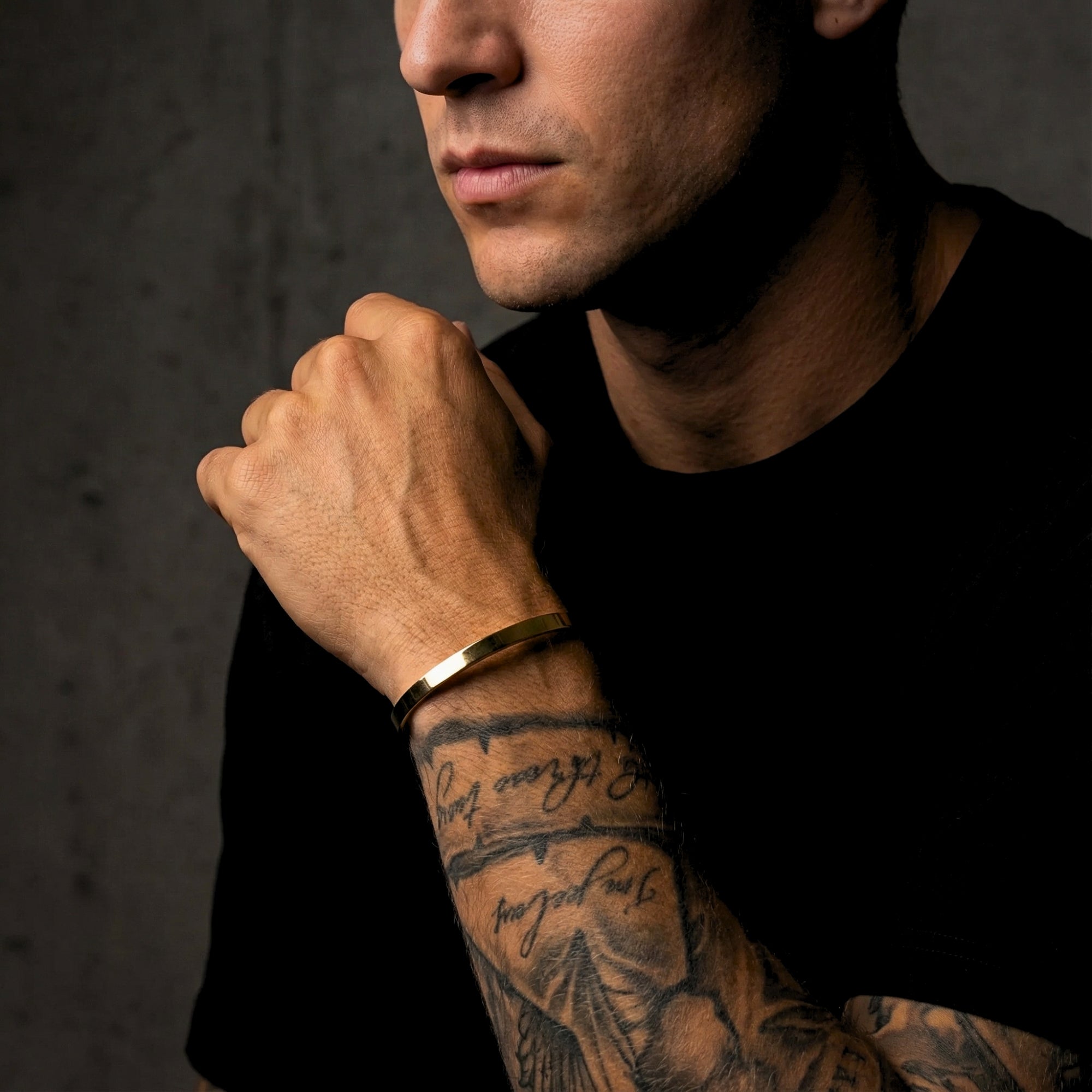 Men's gold stainless steel cuff bracelet worn on a tattooed wrist by a model in a black t-shirt.