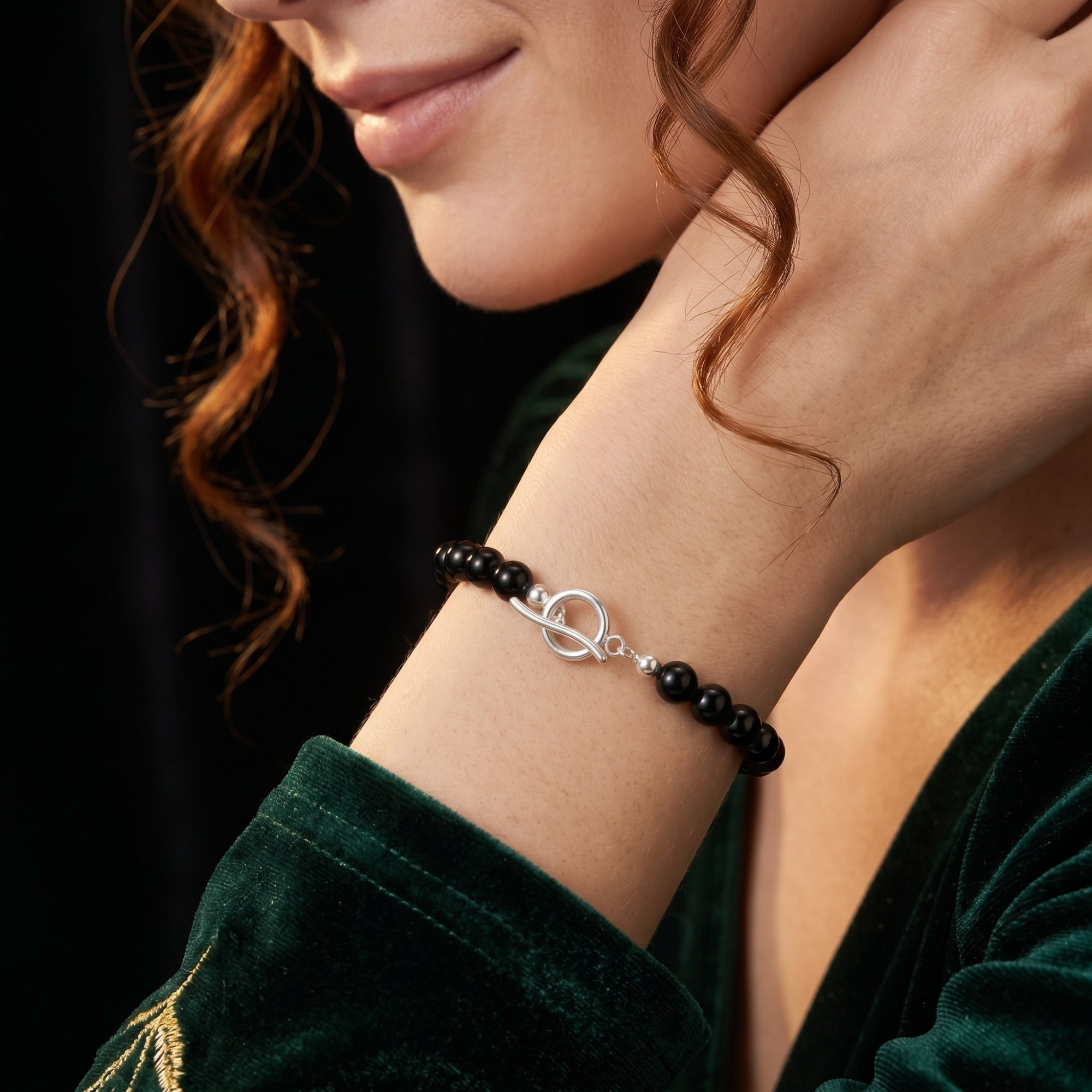 Black onyx bead bracelet with a silver toggle clasp worn on a woman's wrist, paired with a green velvet dress.