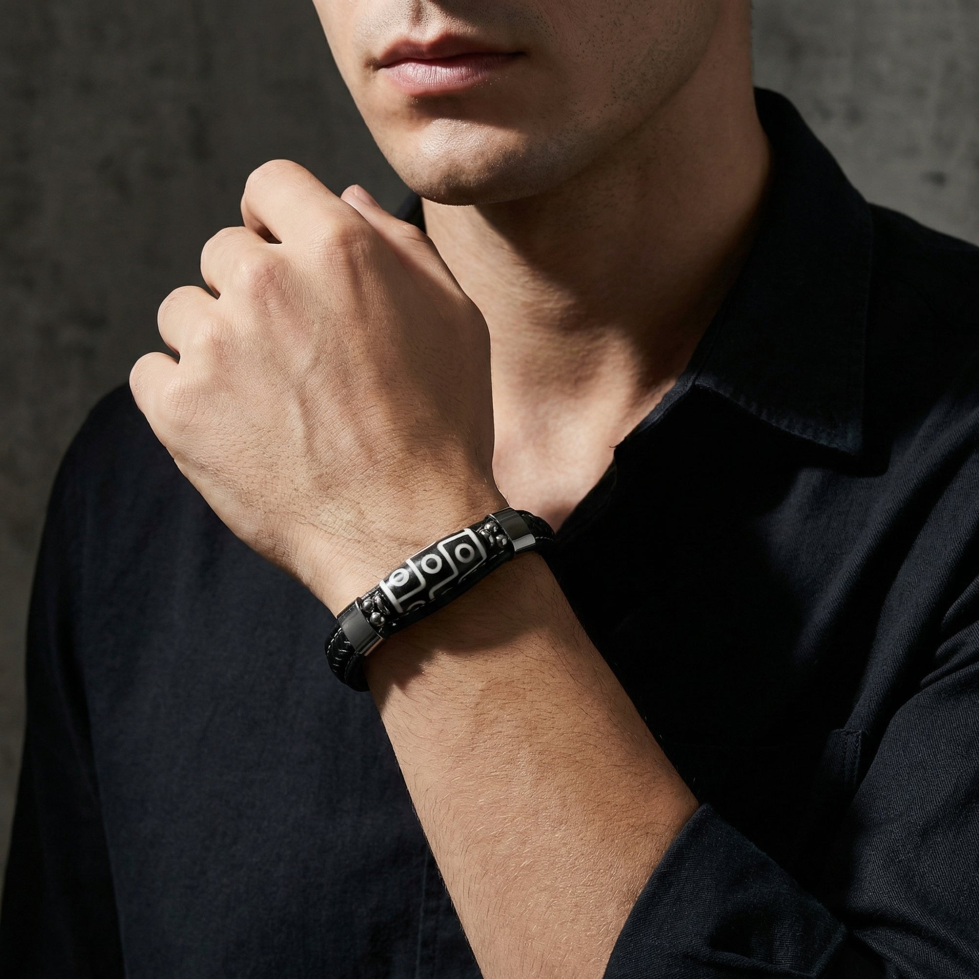 Men's black leather bracelet with a silver-tone 316L stainless steel centerpiece worn on a wrist with a black shirt.