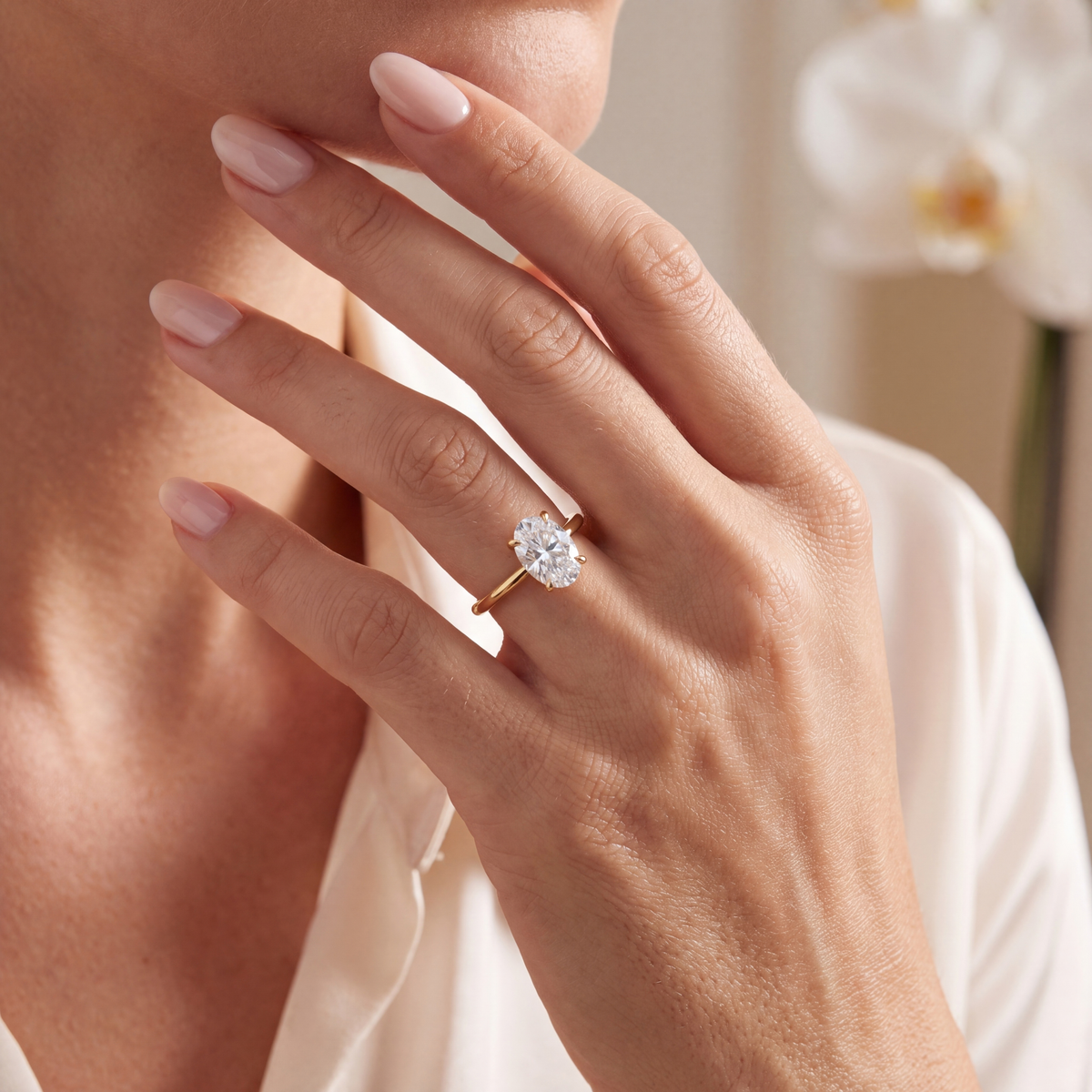 An oval moissanite solitaire ring in yellow gold worn on a hand touching the chin.