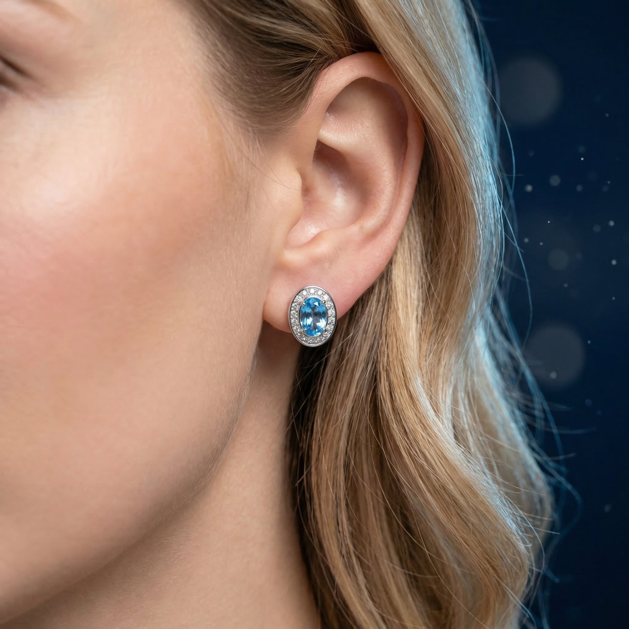 A woman wearing a white gold oval natural blue topaz halo stud earring on her ear.