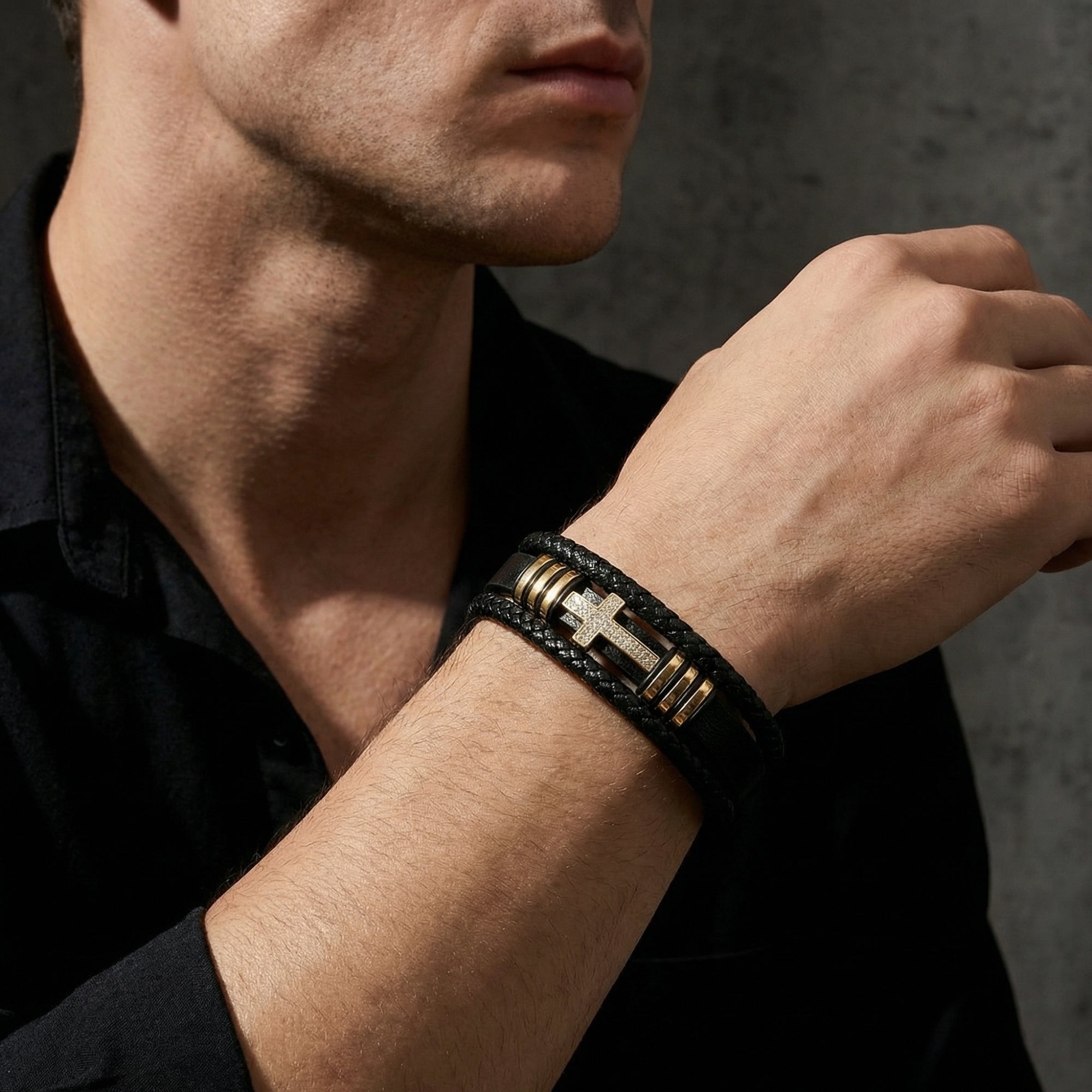 Men's black braided leather bracelet with a gold-finished stainless steel pavé cross, worn on a wrist.