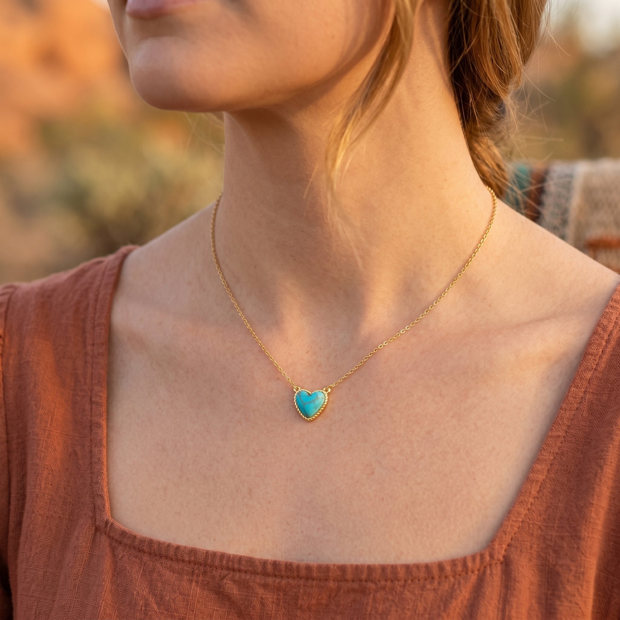 Model wearing the gold Prescott heart necklace with a blue turquoise stone against a soft outdoor desert background.