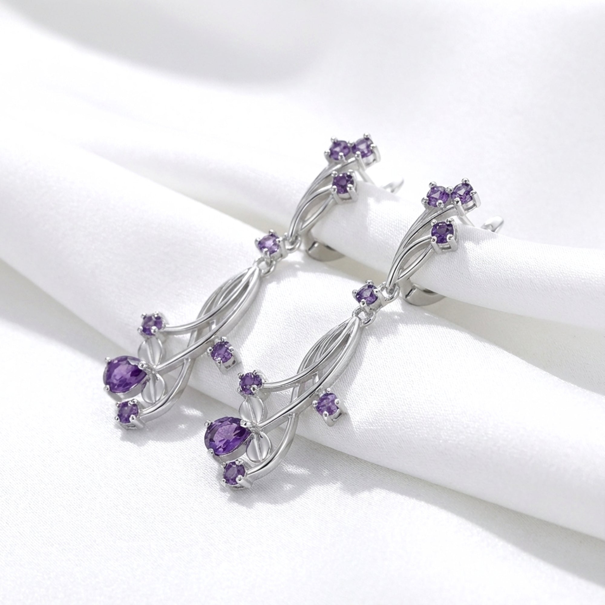 Macro view of S925 sterling silver Lyriel drop earrings with pear-cut simulated amethyst on white fabric.