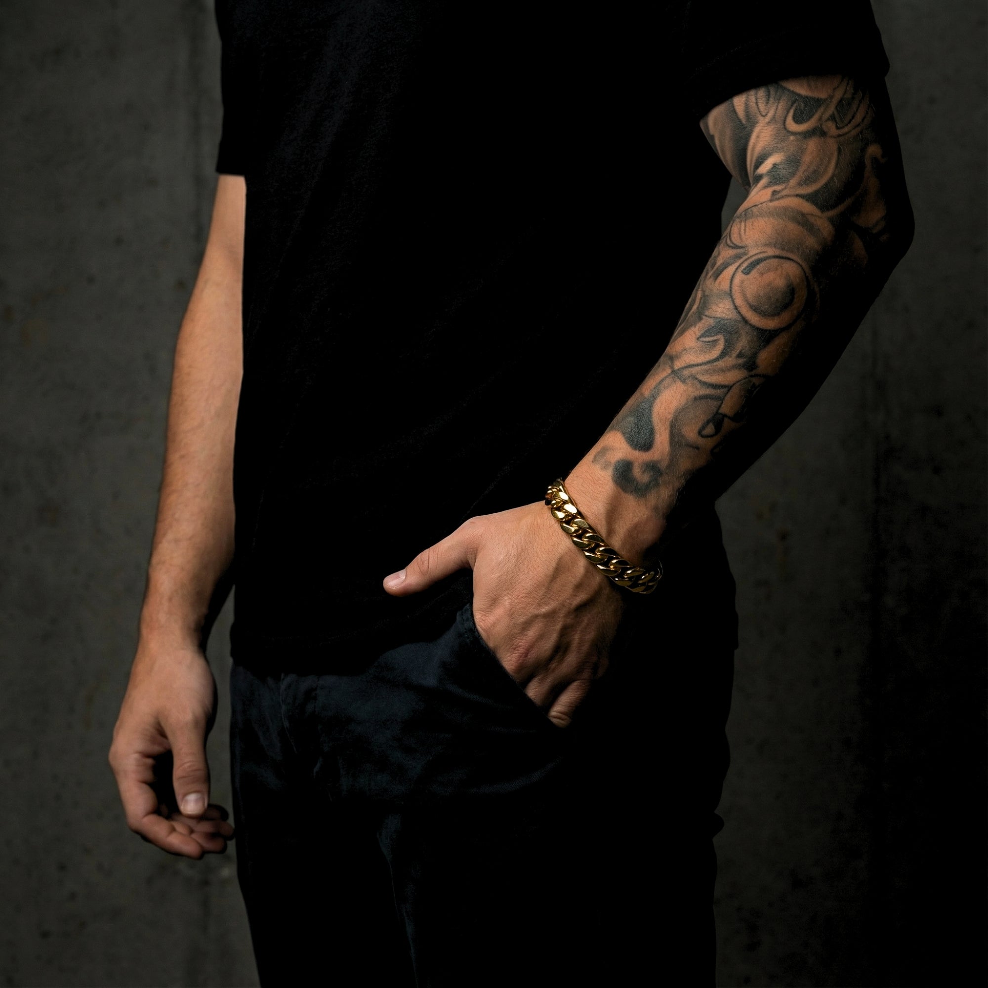 Gold stainless steel Cuban link bracelet worn on a man's tattooed wrist, styled with a black t-shirt.