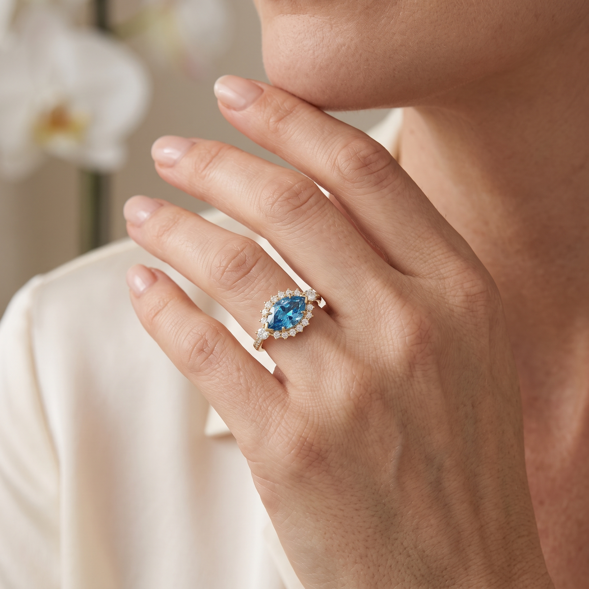 Woman wearing a rose gold marquise blue moissanite halo ring on her hand resting against her chin.