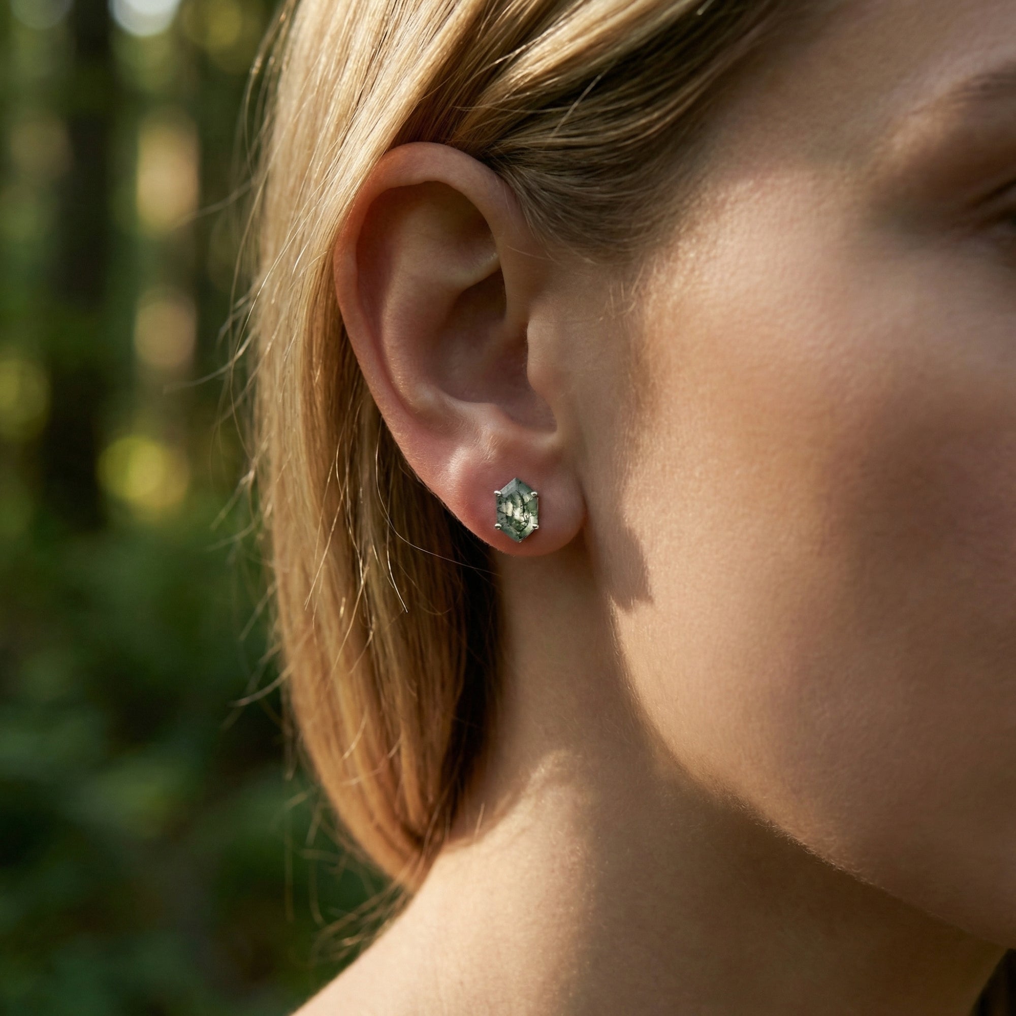 Model wearing a coffin cut natural moss agate stud earring in sterling silver against a forest background.