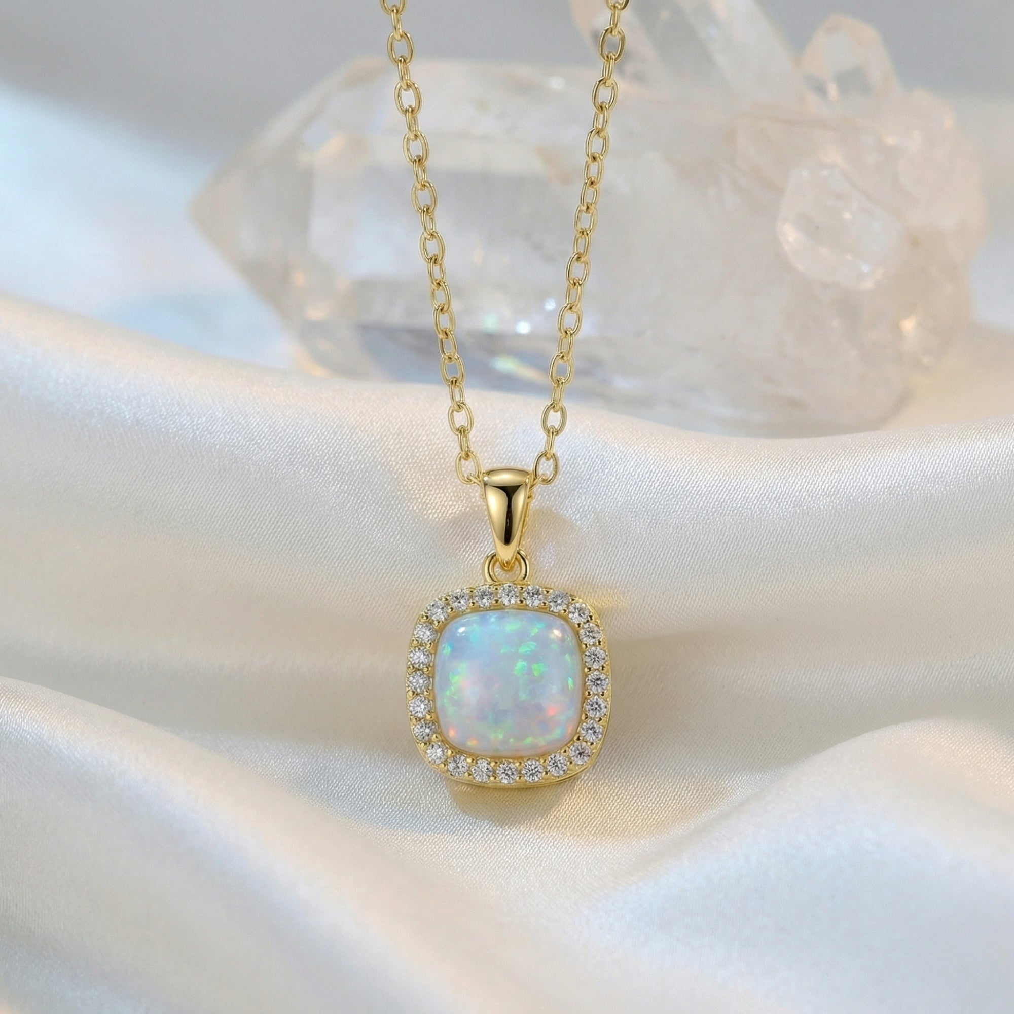 Gold cushion cut white opal halo pendant necklace displayed on white silk fabric with crystal props.