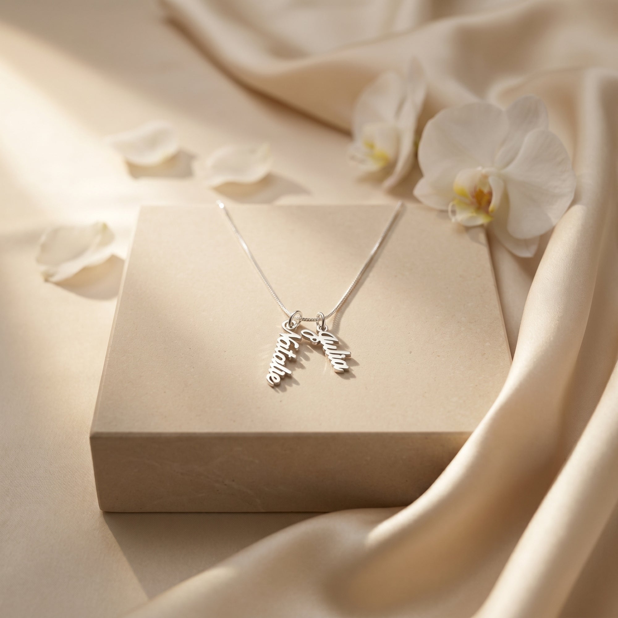Silver name necklace with two vertical pendants on a pedestal with white flowers.