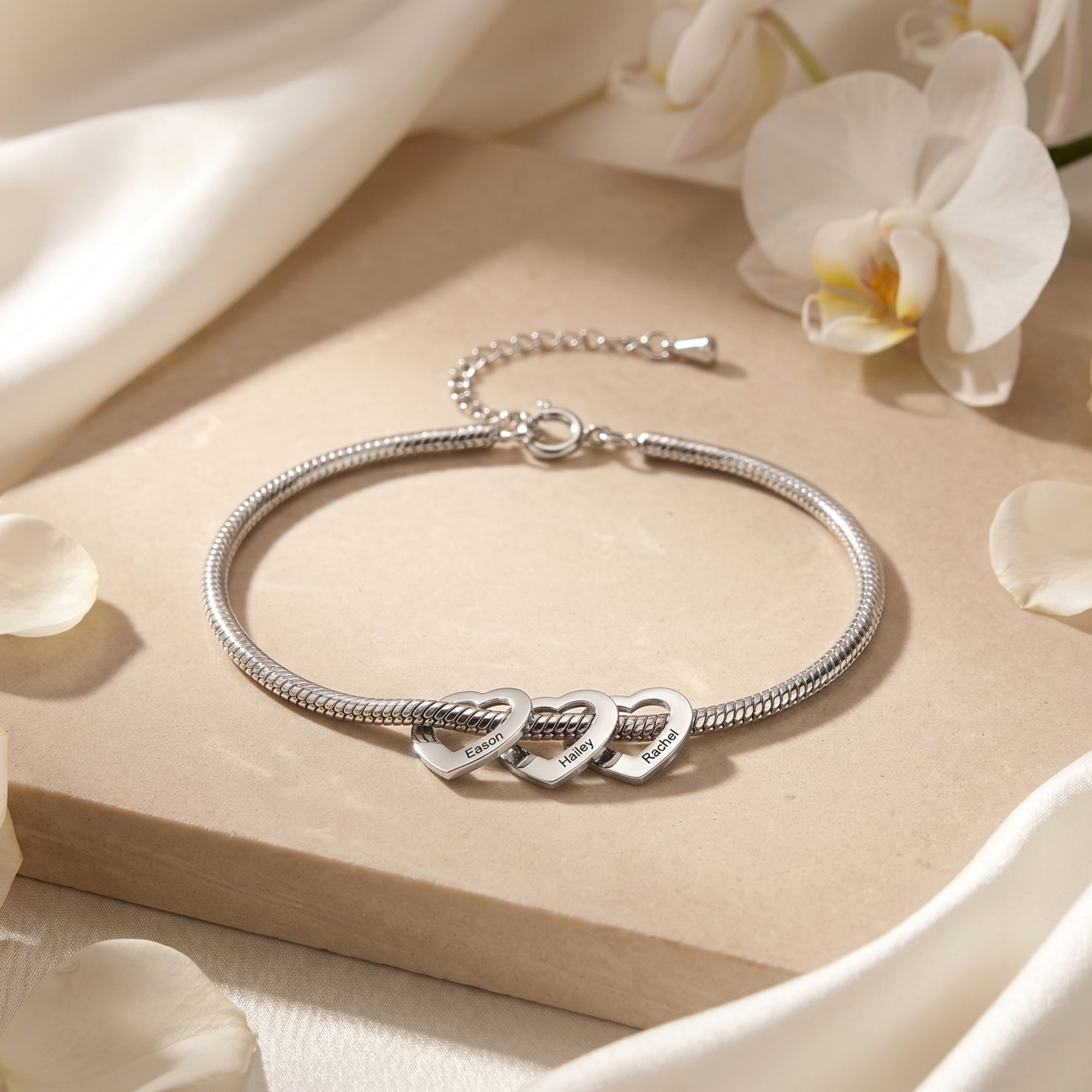 Silver stainless steel snake chain bracelet with three personalized heart charms on a stone surface with flowers.