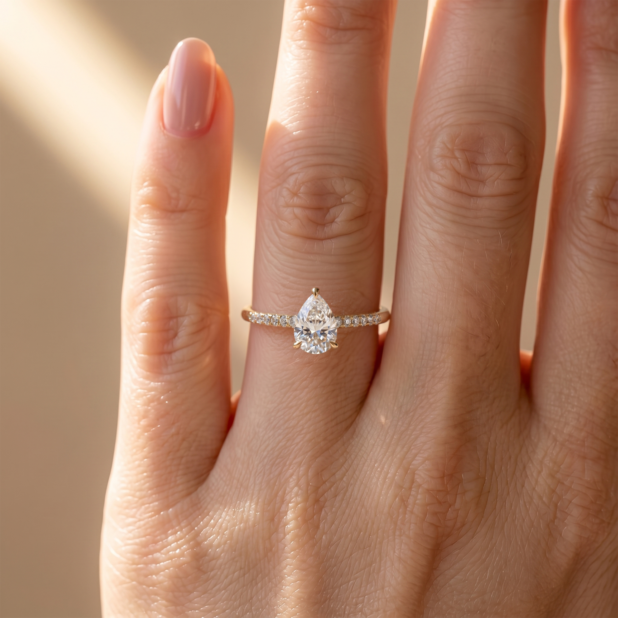 A close-up view of a hand wearing a delicate yellow gold pear-cut moissanite engagement ring with a pave band.