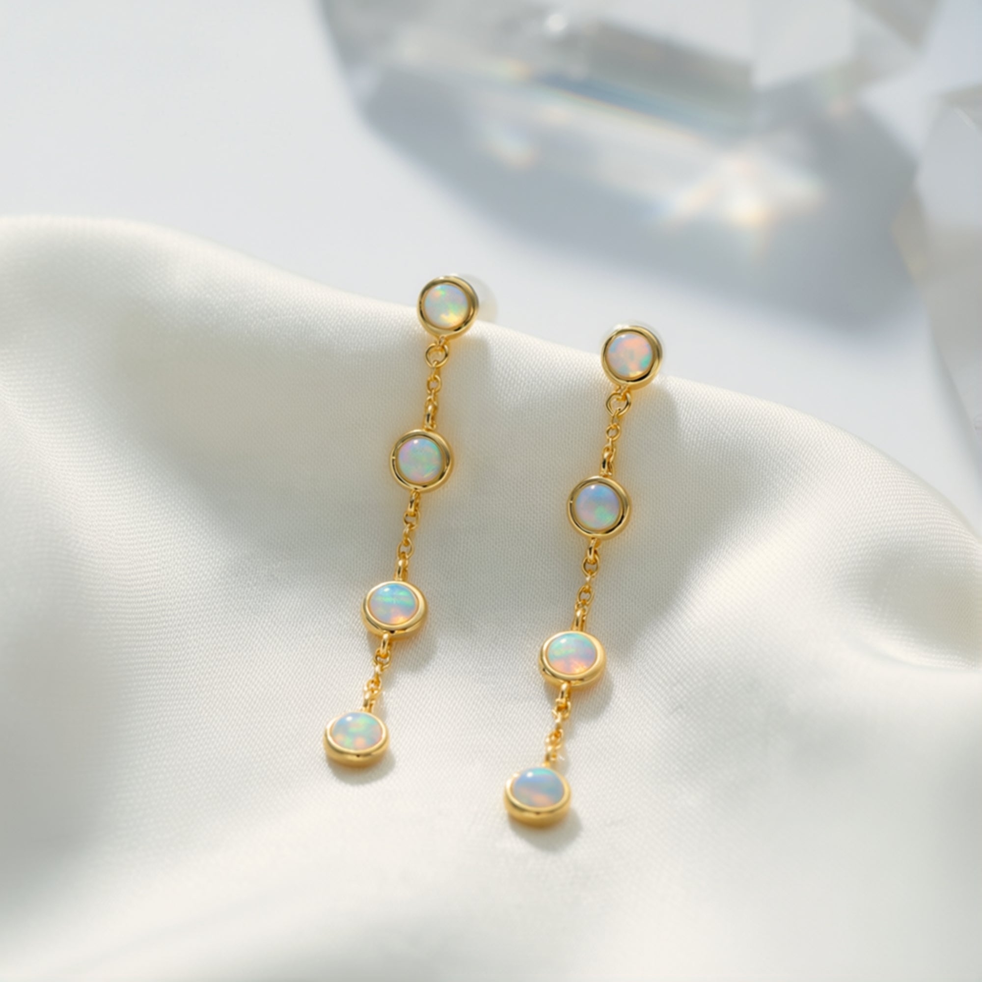 Gold drop earrings featuring a linear cascade of four round white opals on white silk fabric.