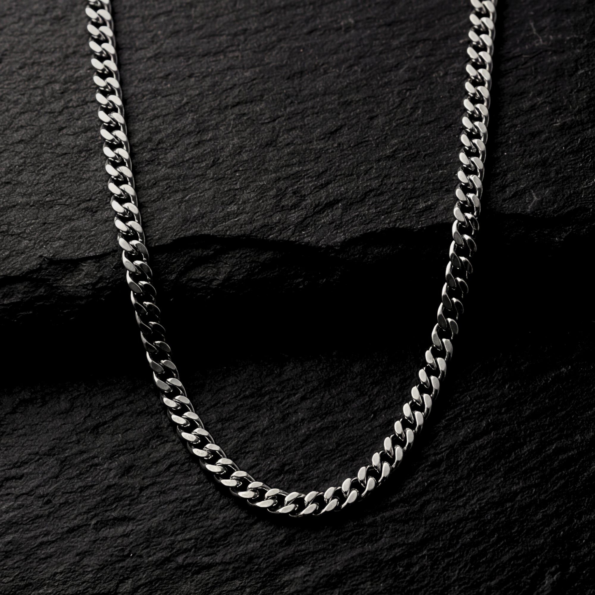 Silver 4mm stainless steel Cuban link chain draped on a dark textured stone background.