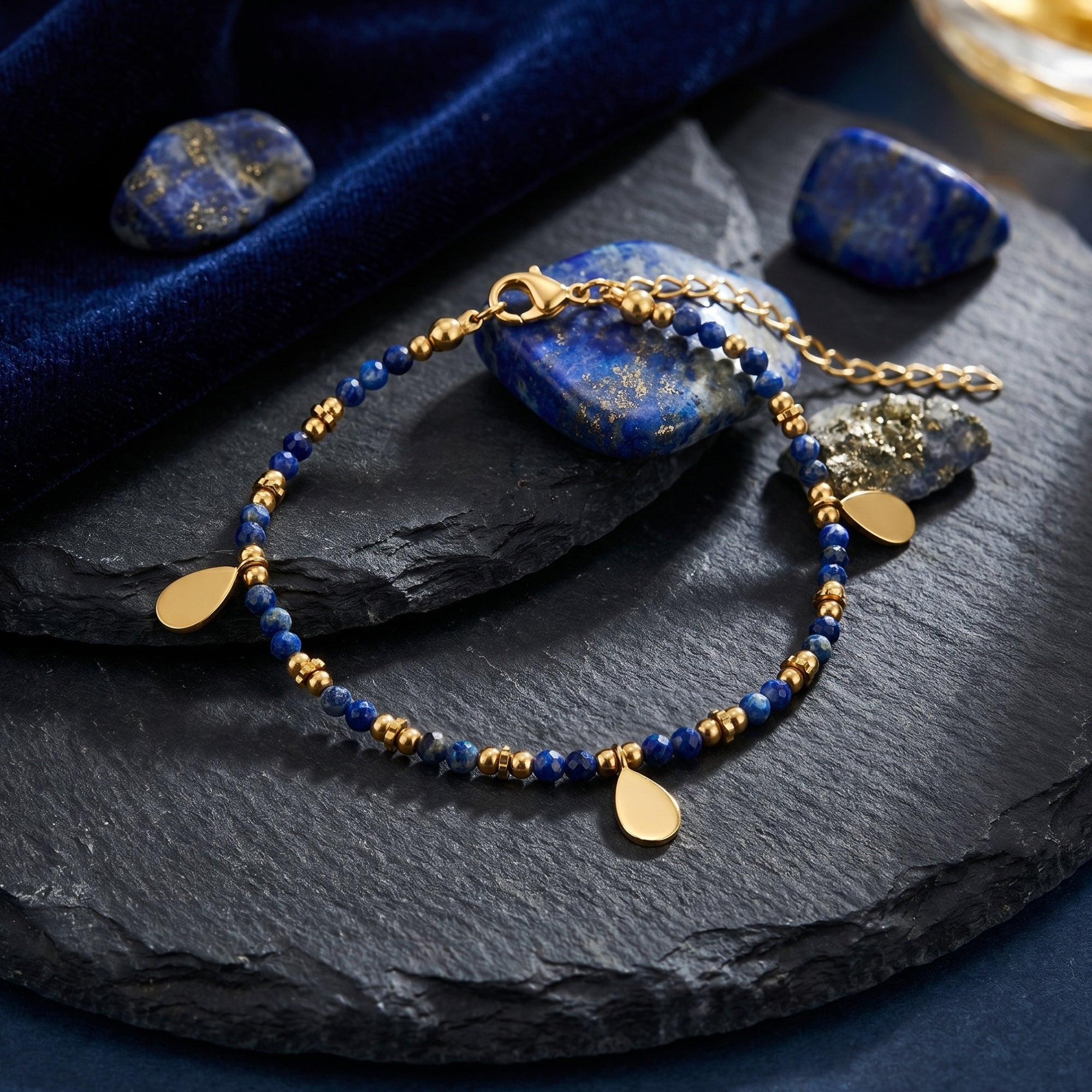 Gold plated faceted 3mm lapis bead bracelet with teardrop charms displayed on slate with raw lapis stones.