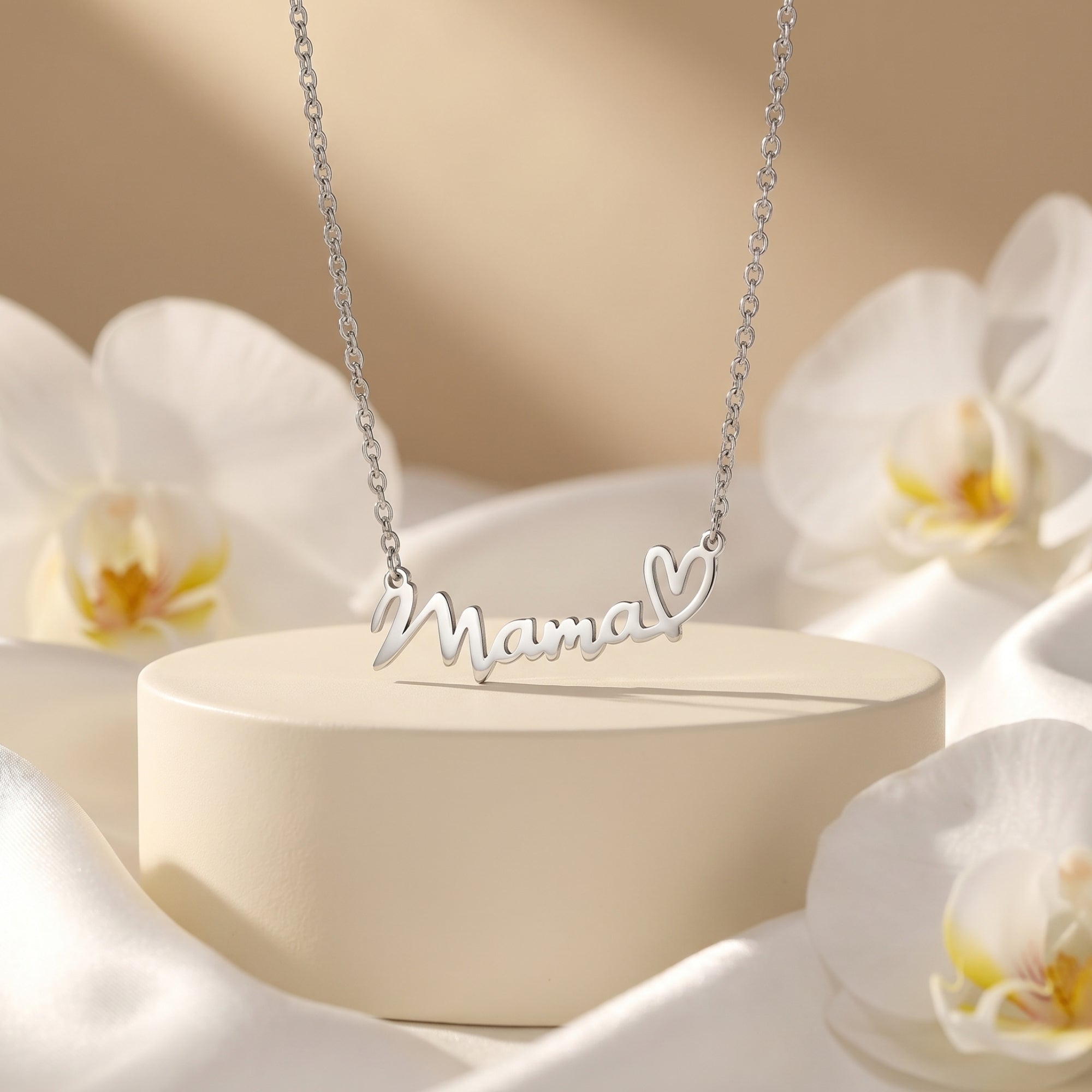 Silver Mama script necklace with heart motif on a pedestal surrounded by white orchid flowers.