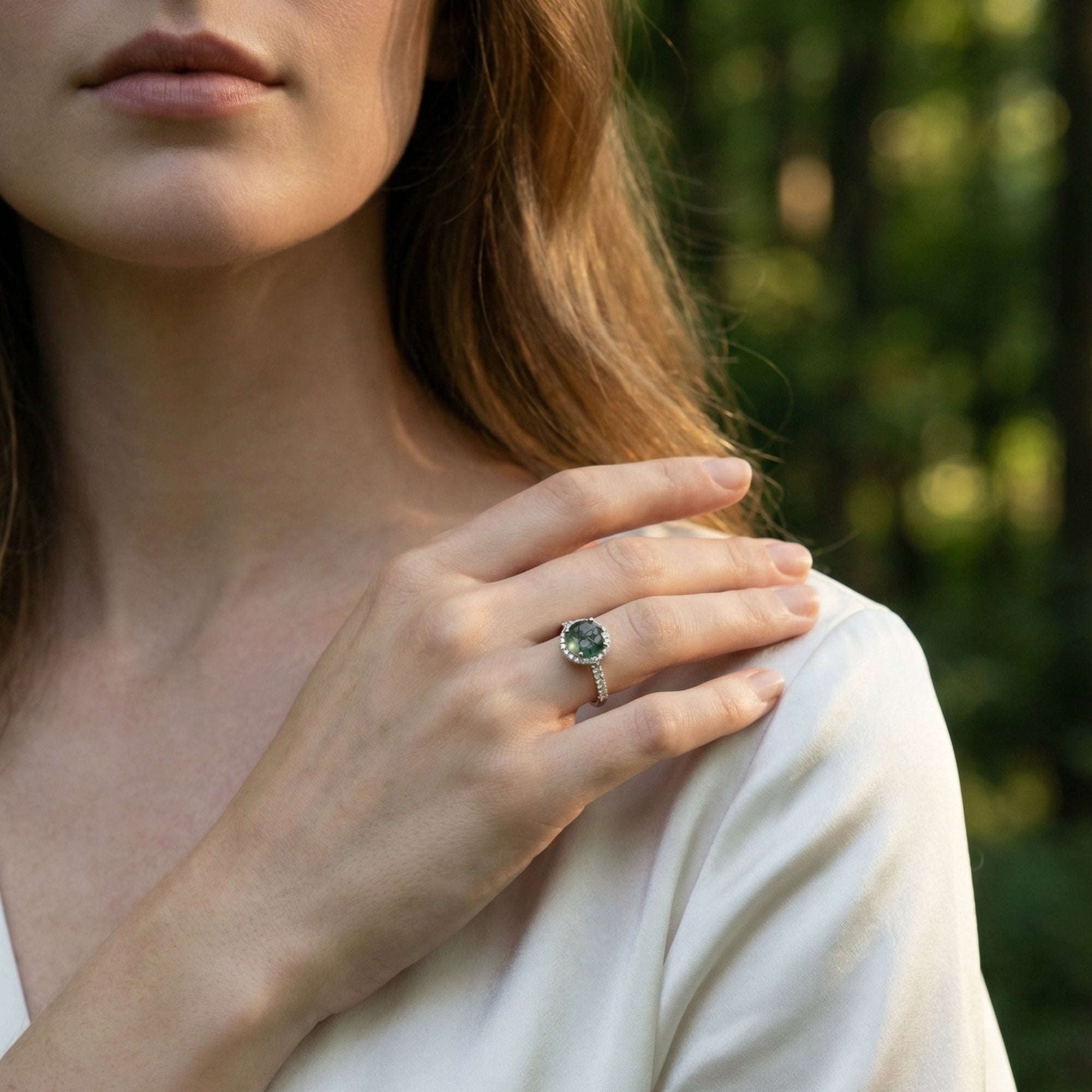 Close-up of a model wearing the natural moss agate and diamond halo ring on her finger against a forest background.