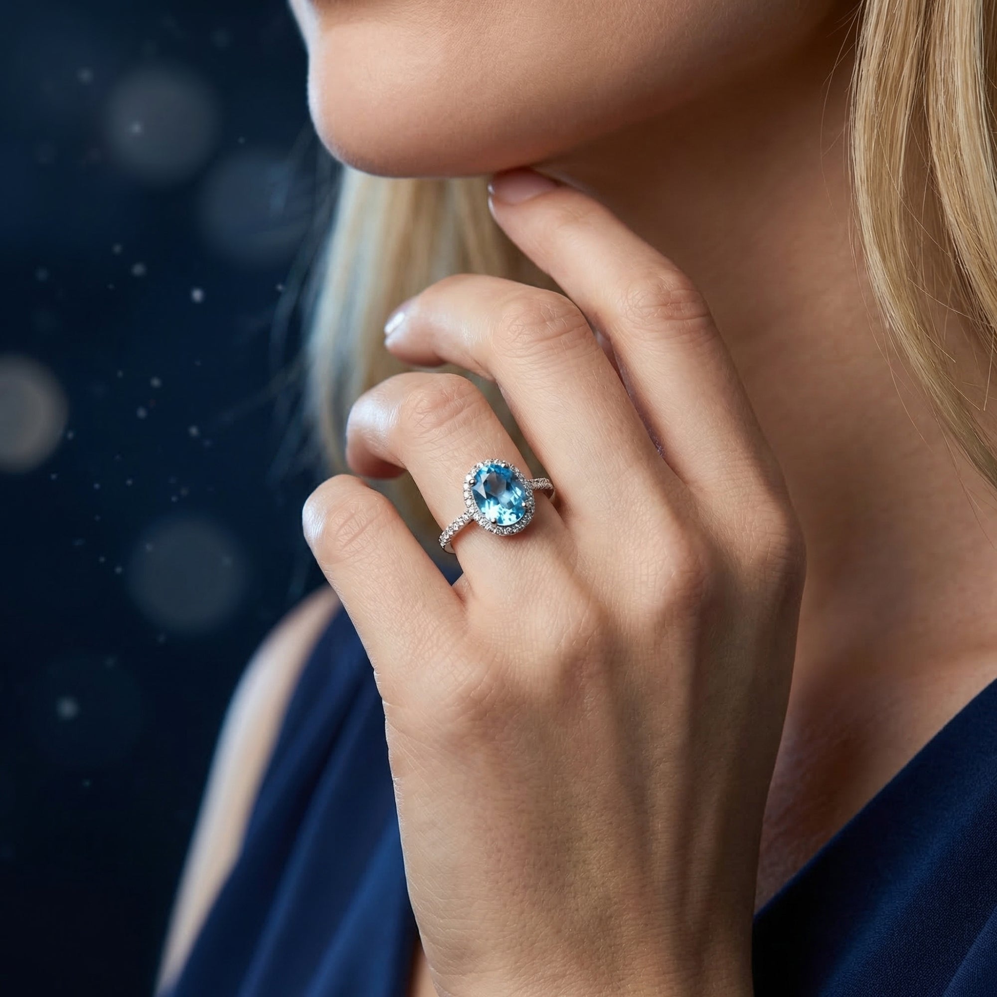 A model wearing the Ariella natural blue topaz sterling silver ring against a dark bokeh background.