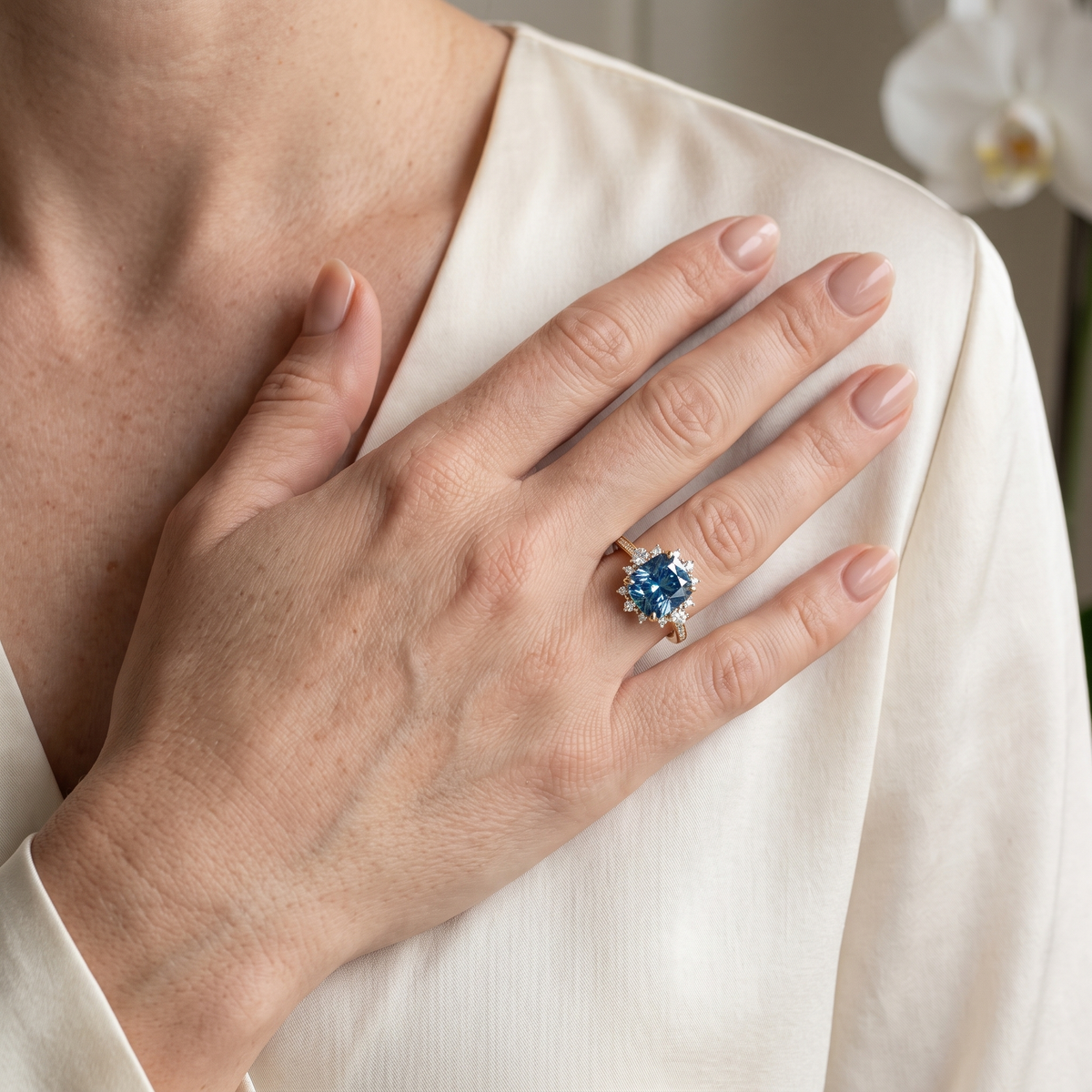 Model wearing a yellow gold cushion-cut blue moissanite ring with hand resting on chest.