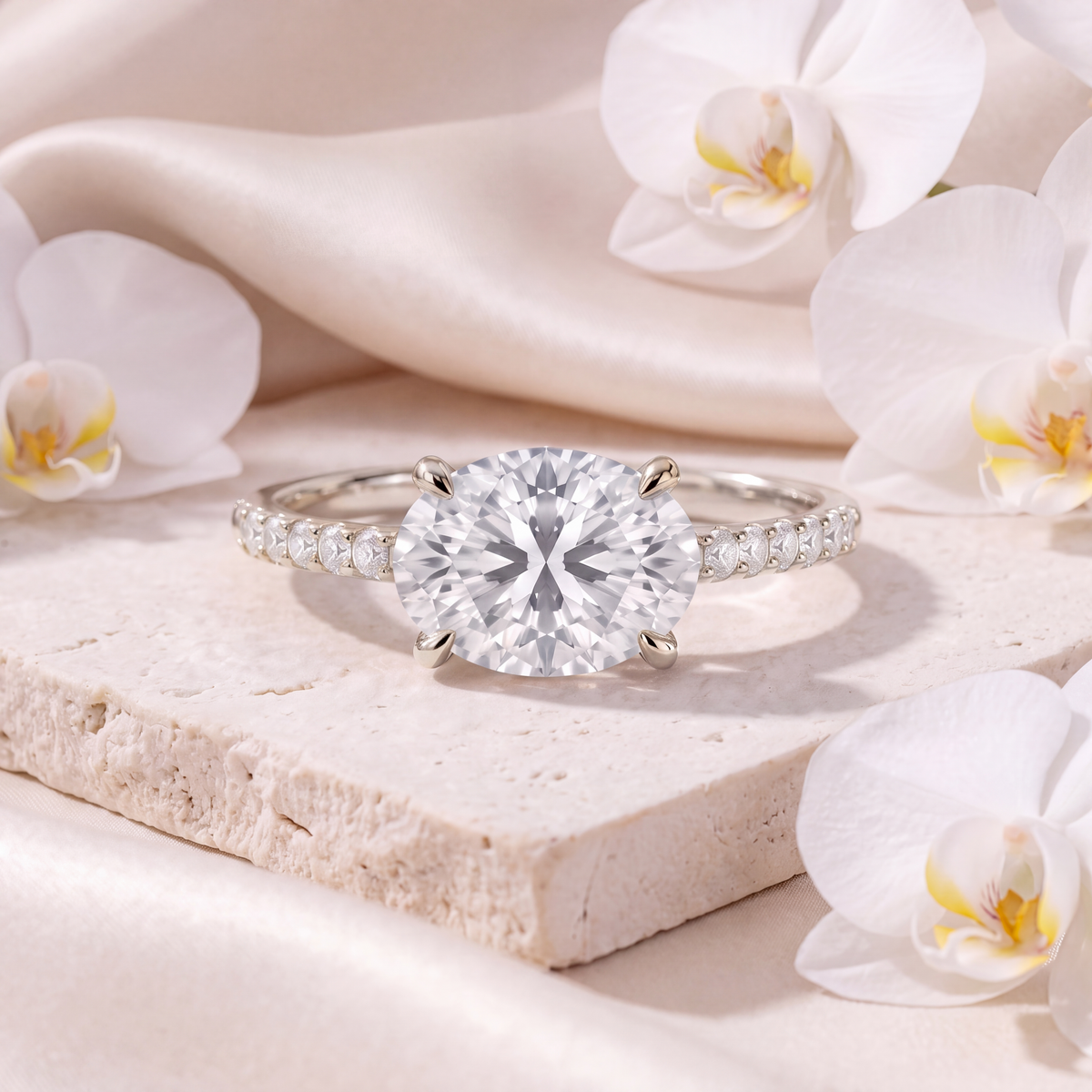 An east-west oval moissanite engagement ring with a pavé band in white gold, presented on travertine stone.