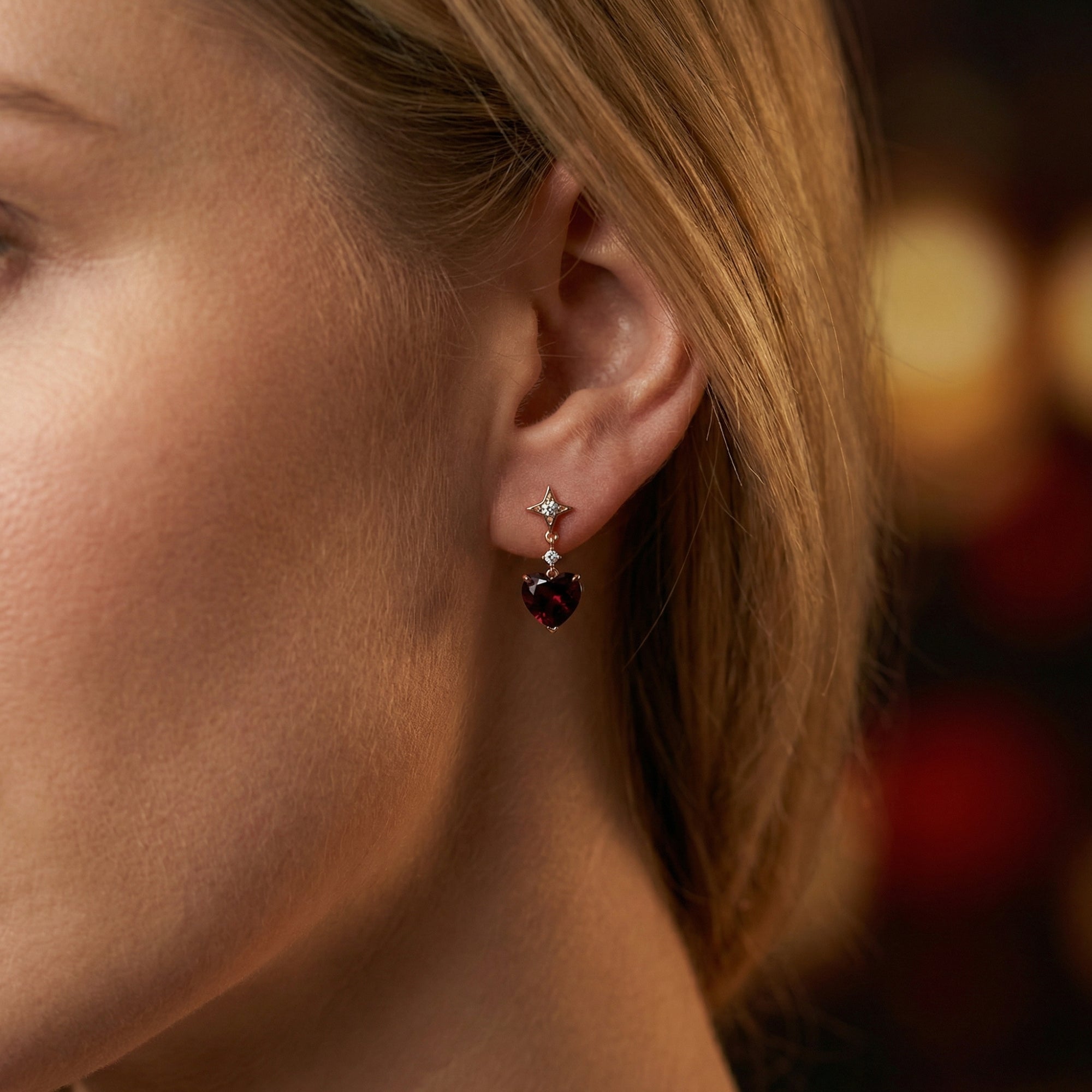 Close-up of a rose gold heart cut garnet drop earring worn on a woman's ear with a soft blurred background.