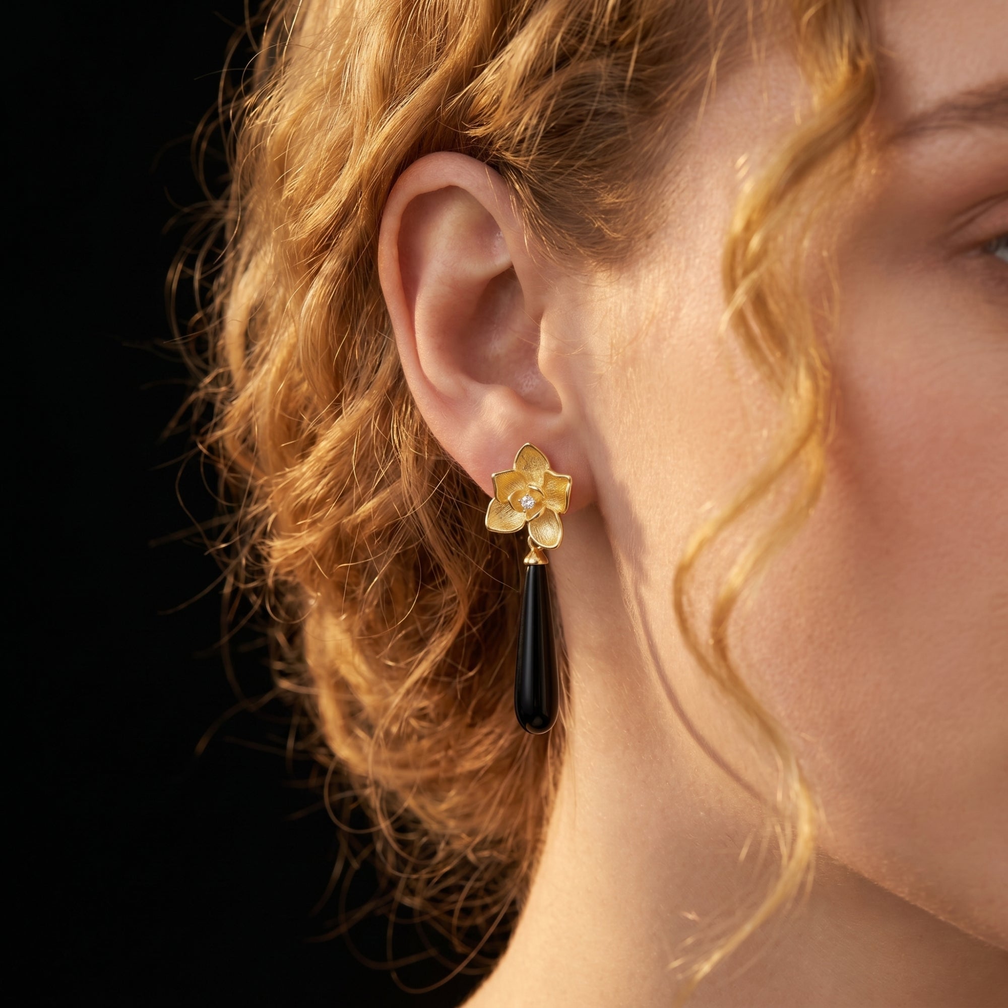 Close-up of an 18K gold plated Calla floral drop earring with an elongated black onyx stone on a model's ear.