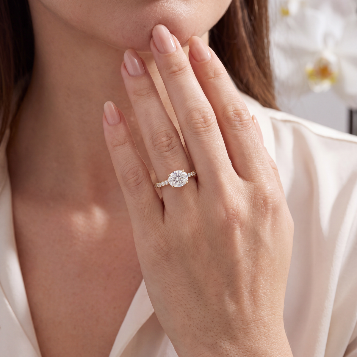 A gold moissanite engagement ring worn on a hand posed near the chin.
