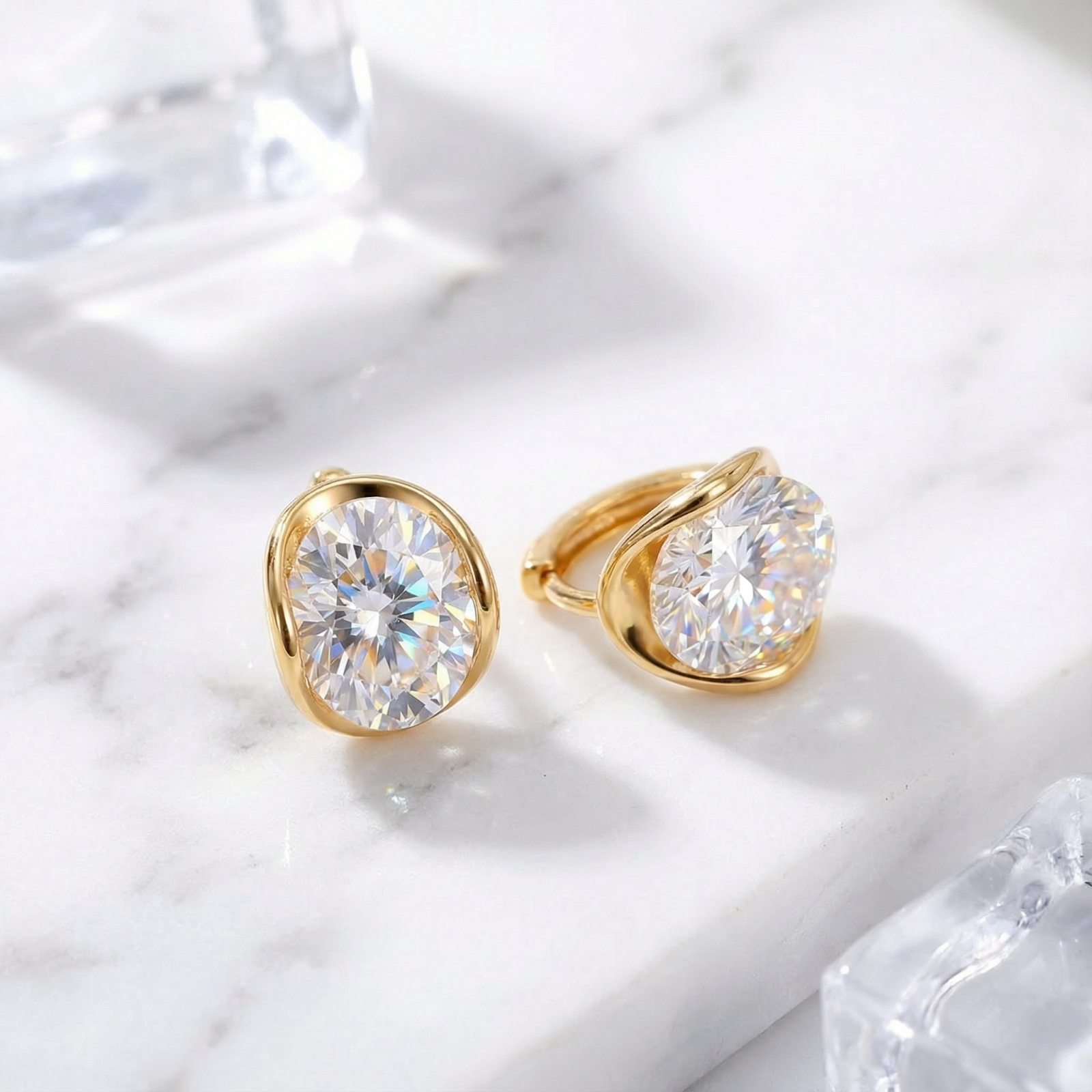 Pair of 18K gold vermeil oval cut zirconia stud earrings on a white marble surface with ice props.