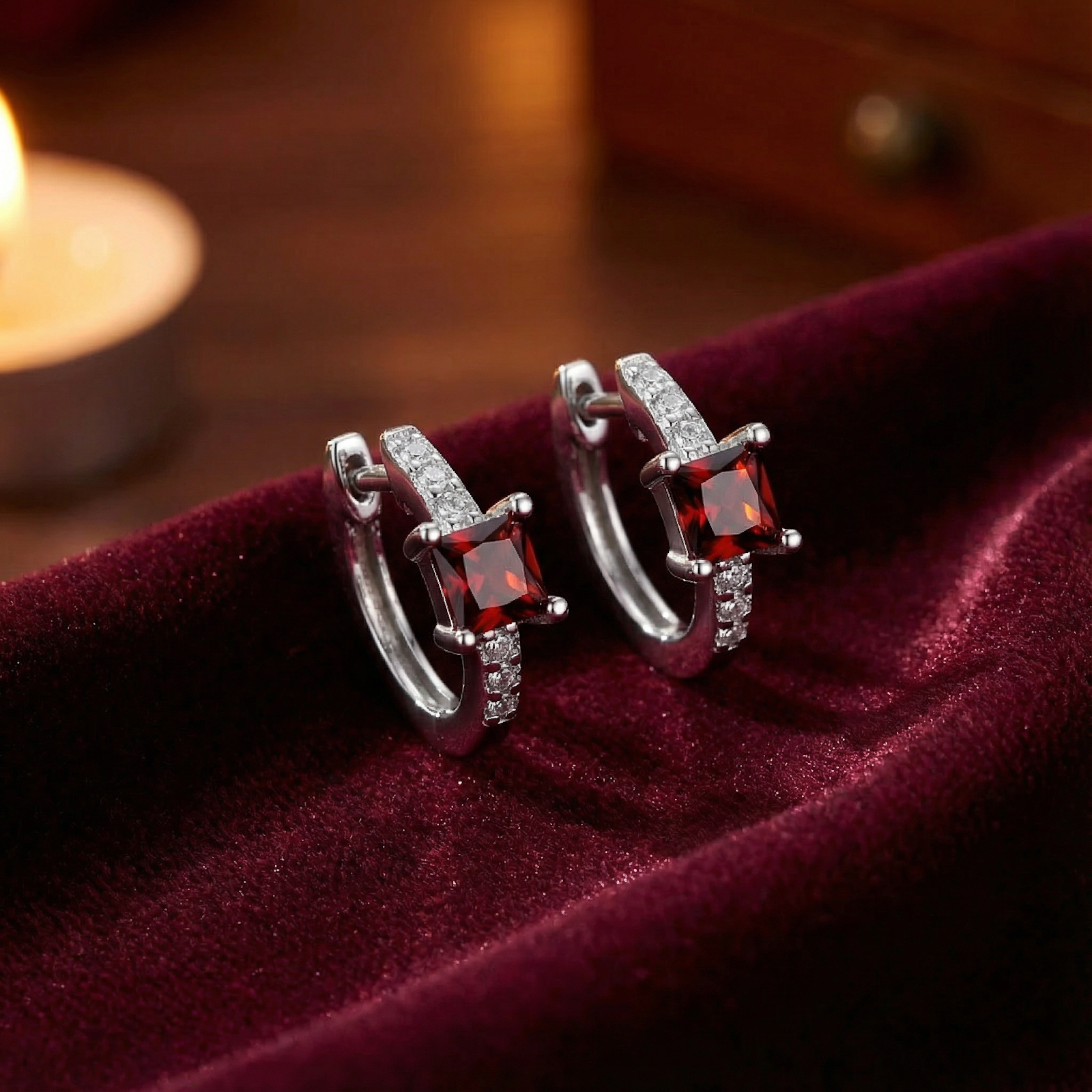 Square cut red garnet huggie earrings in sterling silver resting on red velvet with a warm candle glow.