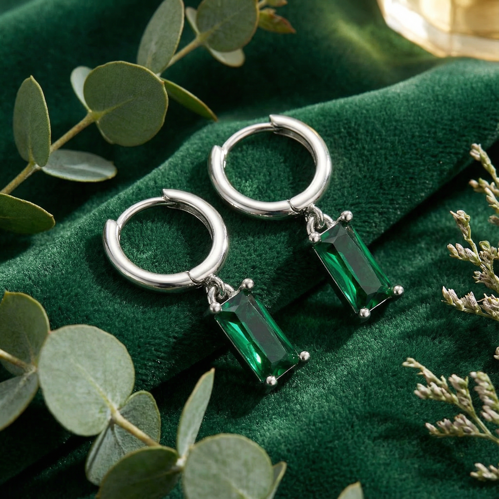 Sterling silver hoop earrings with emerald-cut simulated emerald drops on green velvet fabric with greenery.