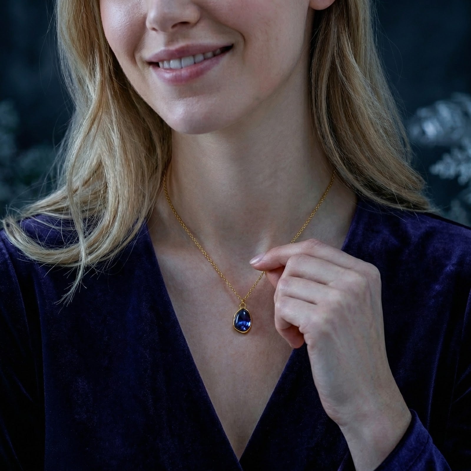 Woman wearing the gold-finished teardrop necklace with a blue-violet simulated tanzanite, hand touching the chain.