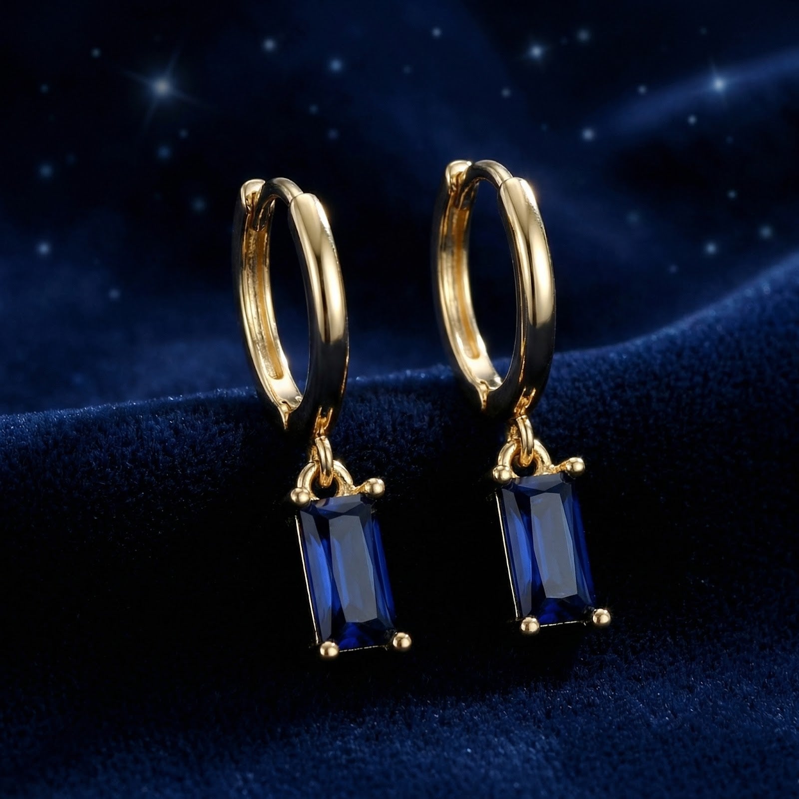 Pair of gold vermeil hoop earrings with emerald-cut blue sapphire charms standing on blue velvet.