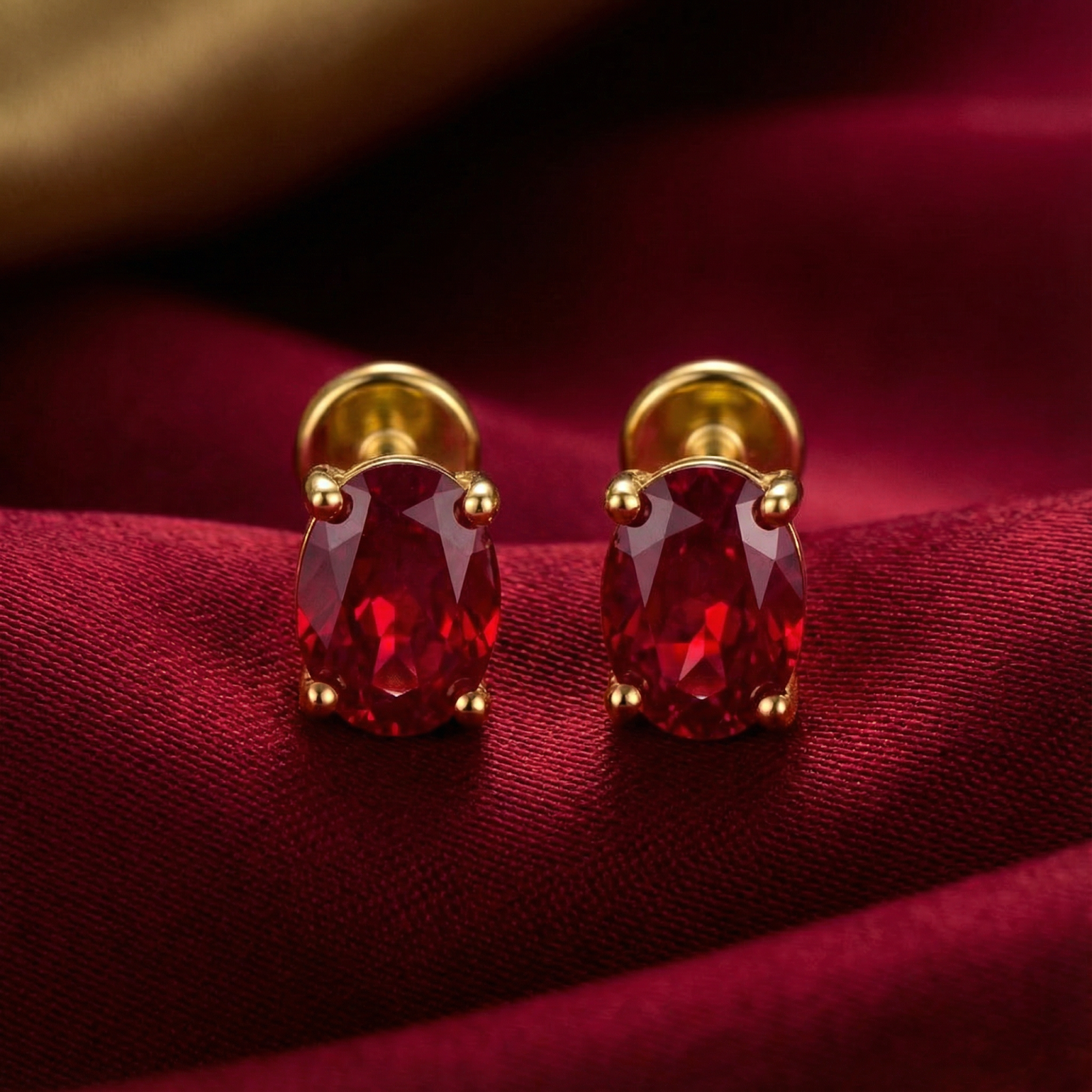 Front view of oval cut simulated ruby earrings in 18K gold vermeil settings on red satin fabric.