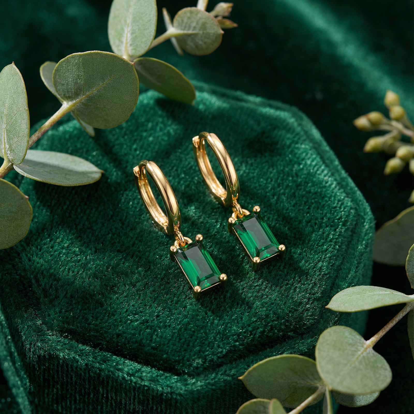 Macro view of gold vermeil drop earrings featuring precision-cut emerald-cut simulated emeralds on green velvet.