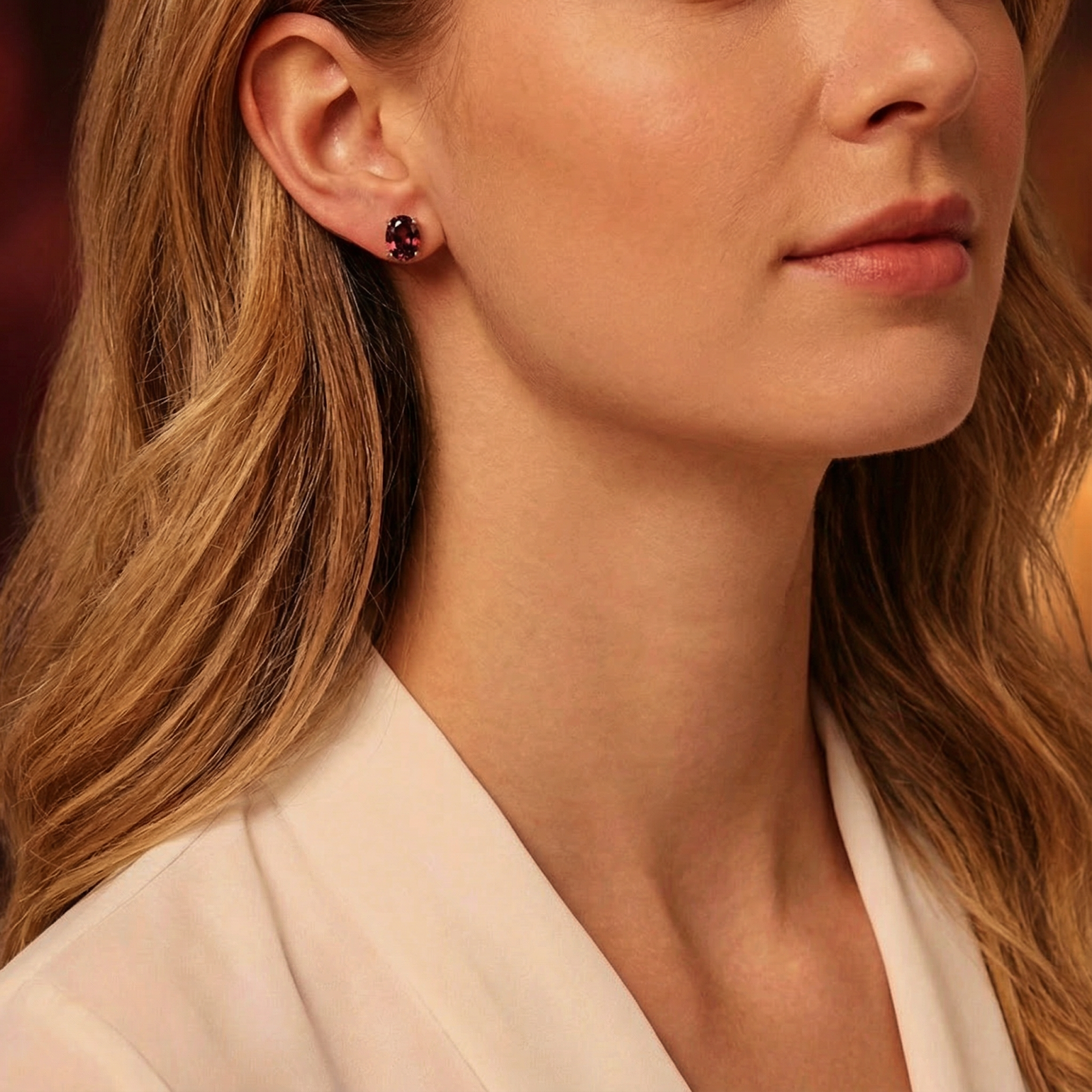A woman wearing the Aderyn oval cut simulated garnet stud earrings in sterling silver with a white blouse.
