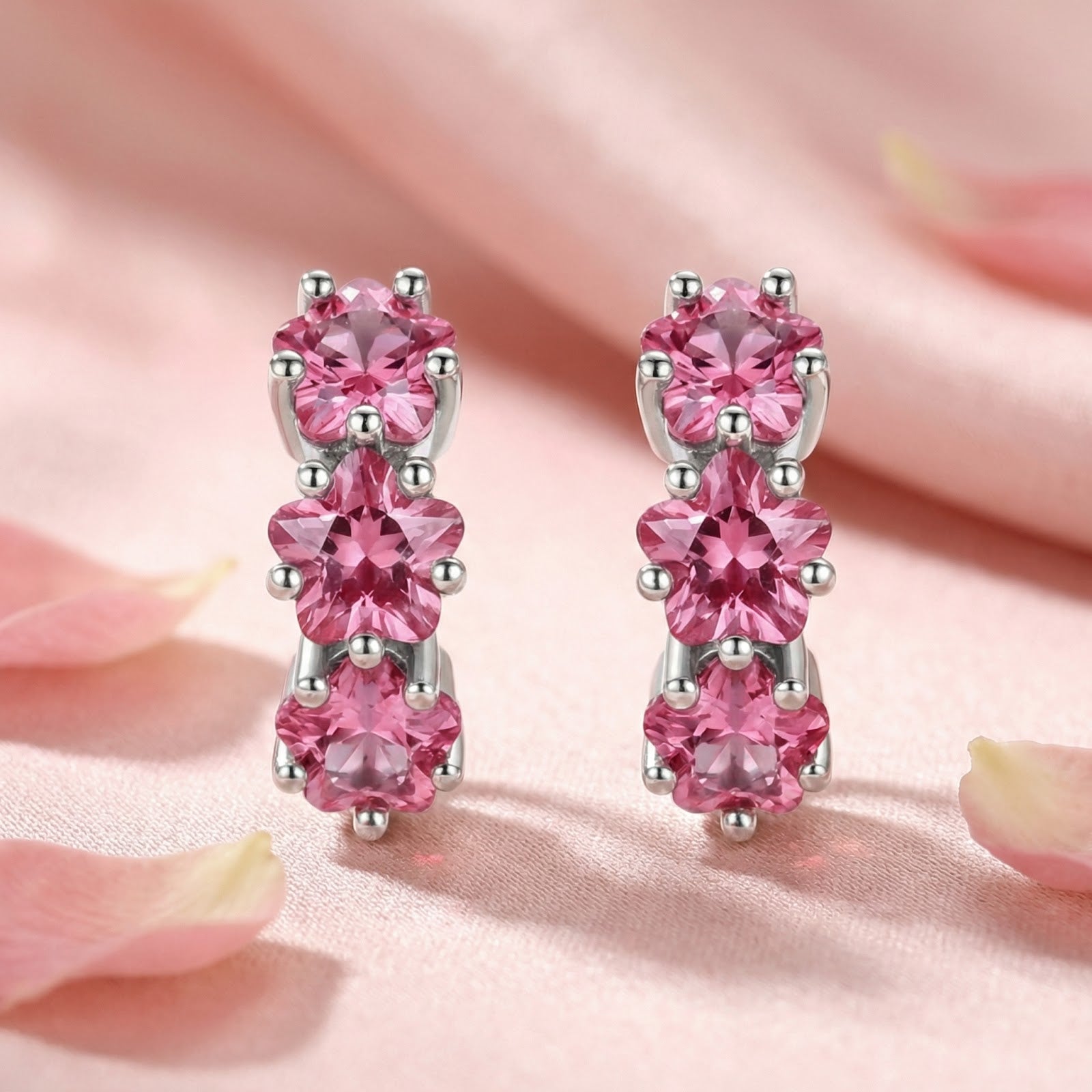 Front view of silver star huggie earrings with three star-cut pink tourmaline stones on a pink fabric background.