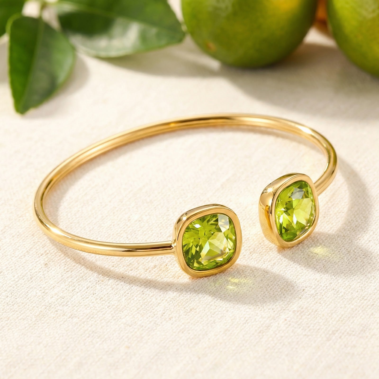 Gold-finished stainless steel Zelina bangle with cushion-cut simulated peridots on a white surface with limes.