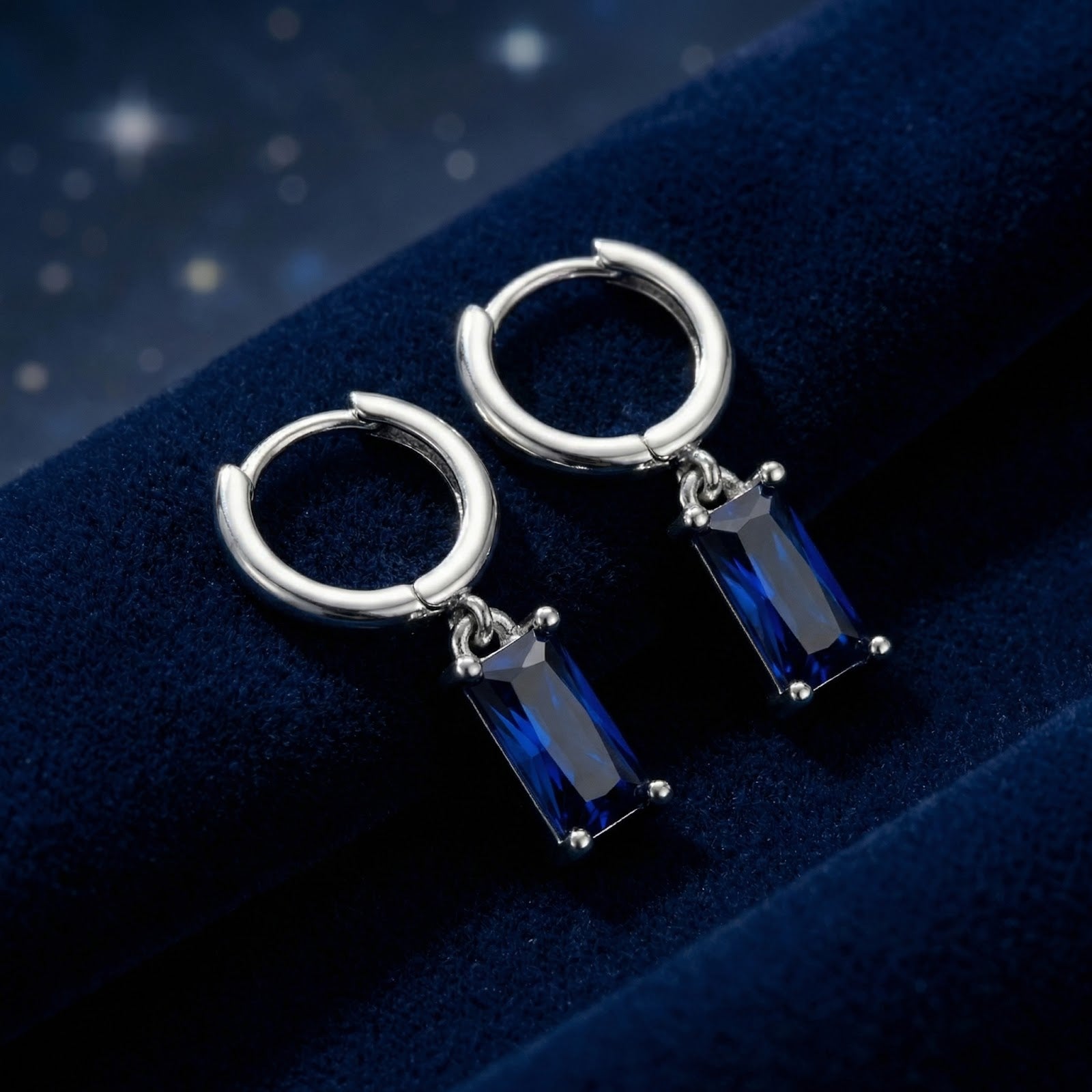 Sterling silver hoop earrings with emerald-cut simulated blue sapphire charms on a dark velvet background.