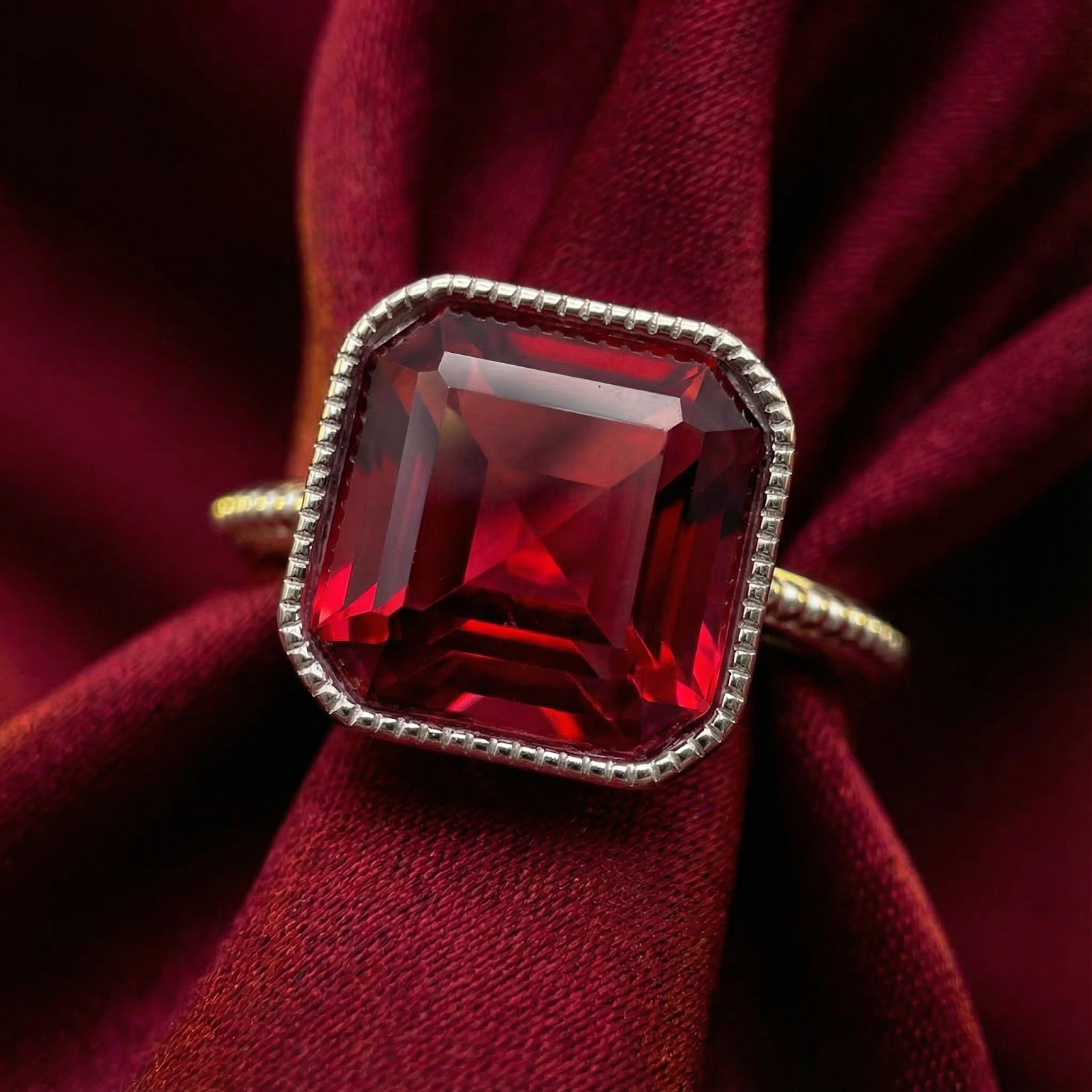 Close-up of a square-cut simulated ruby in a textured S925 sterling silver setting on red fabric.